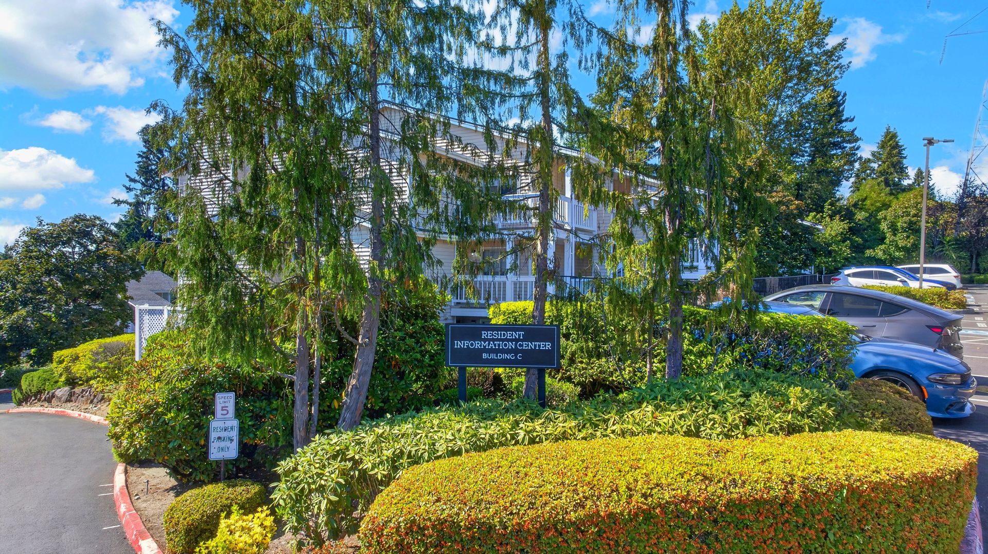 Landscaped office park entrance with trees, hedges, and a parking lot sign beside parked cars