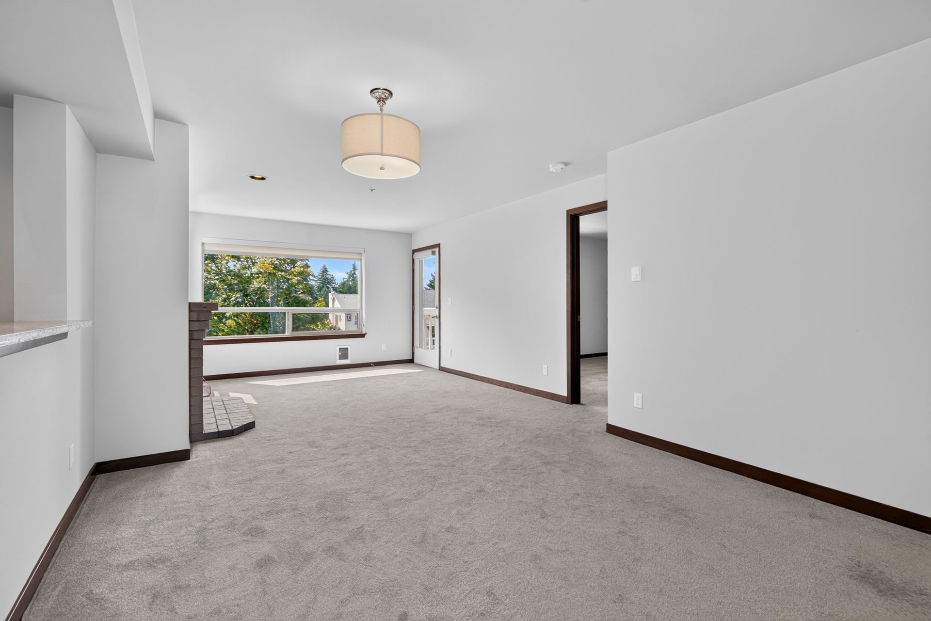 Empty carpeted living room with white walls, dark trim, and large windows overlooking greenery