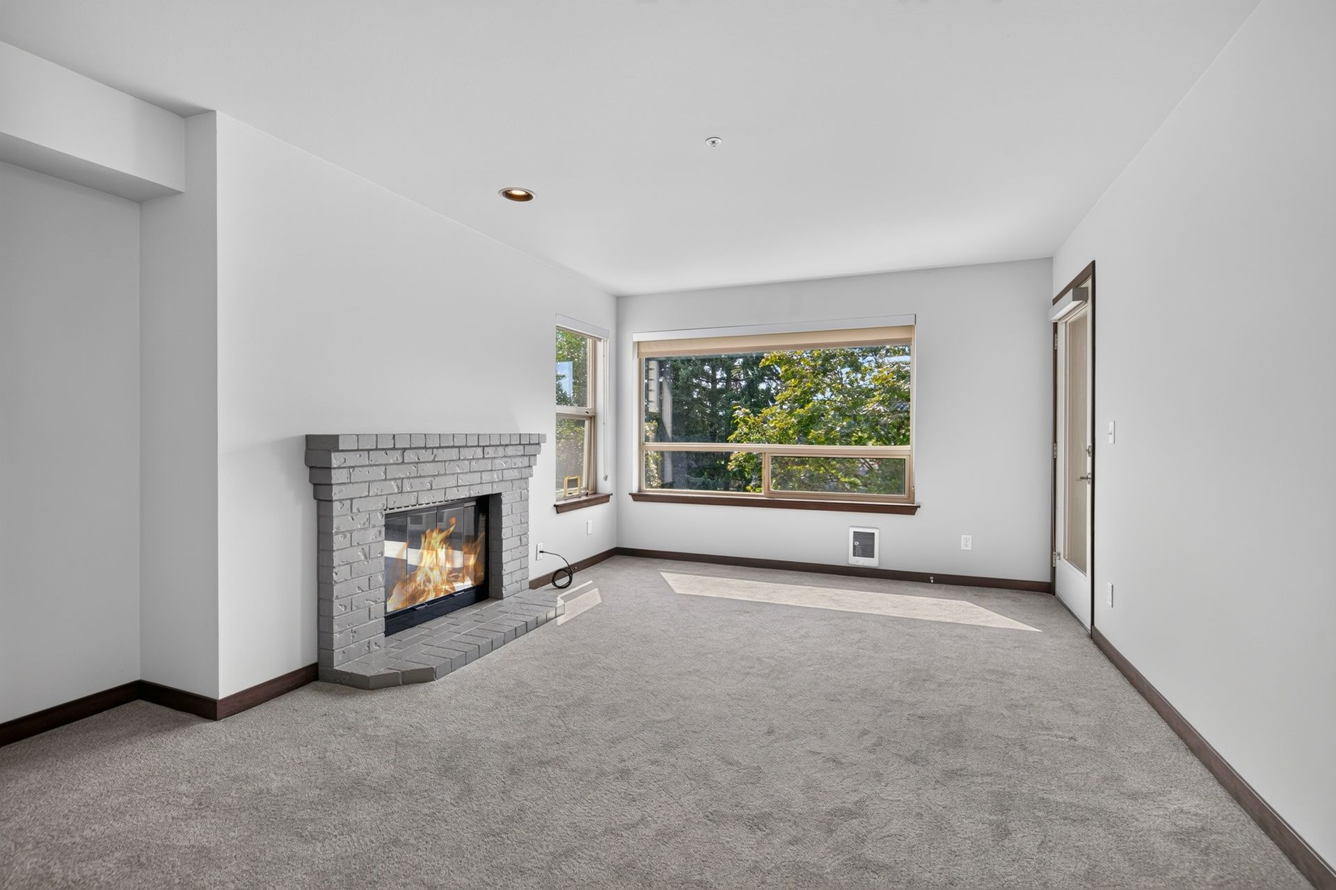 Empty living room with gray carpet, white walls, a fireplace, and a large window overlooking trees