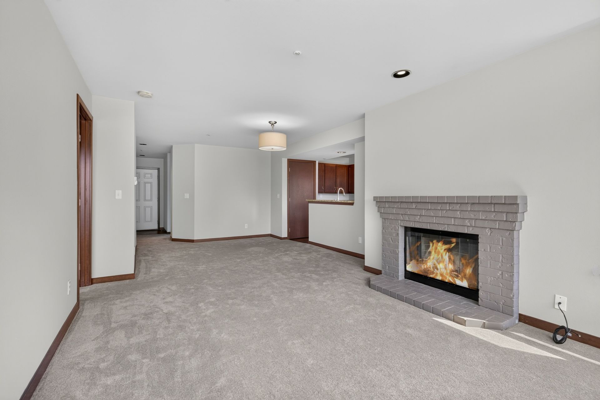 Empty living room with gray carpet, white walls, and a lit fireplace on the right