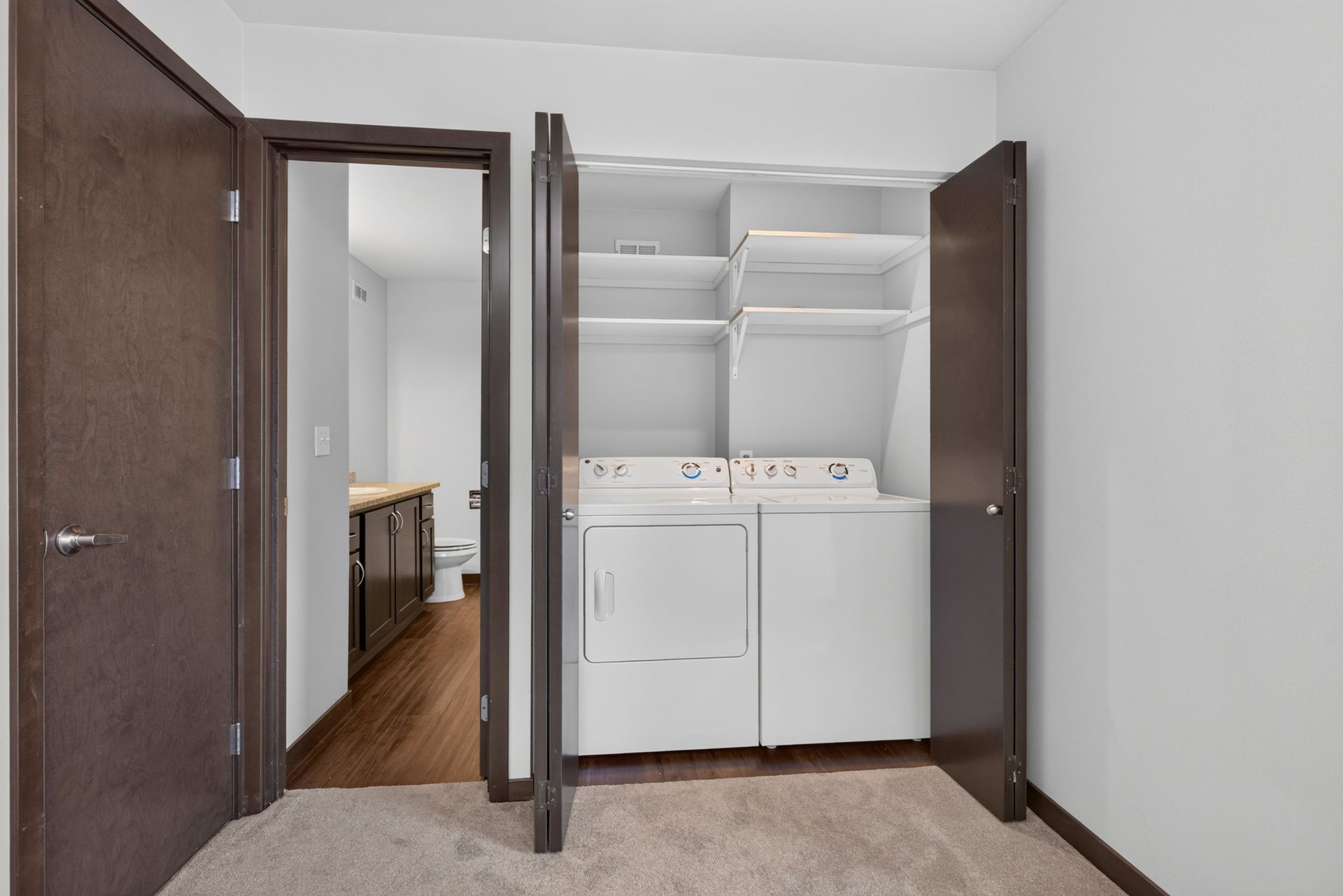 Open closet with white shelves and washer-dryer beside a doorway in a carpeted room