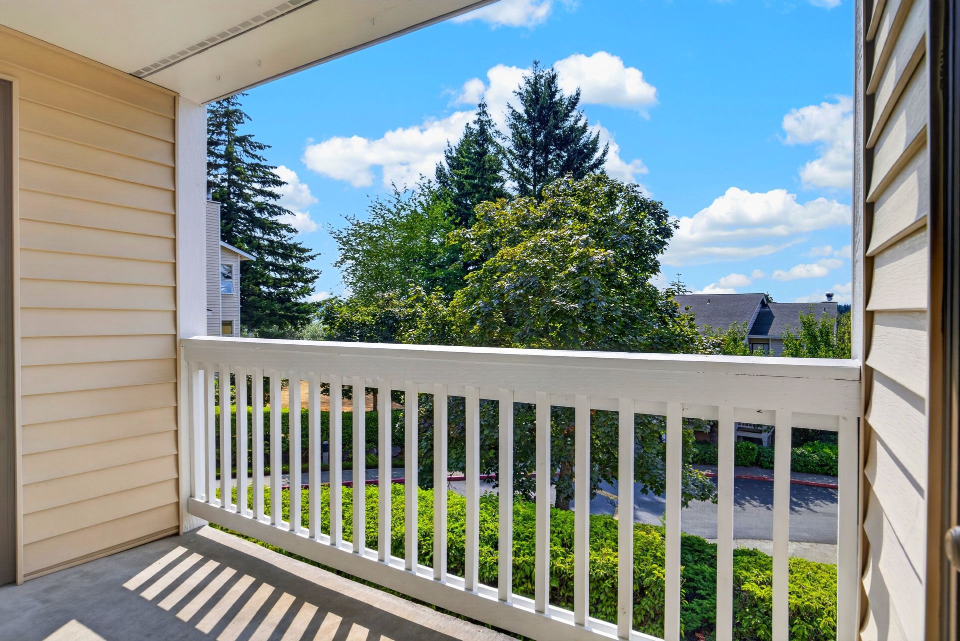 Balcony view with white railing, green trees, blue sky, and neighboring houses in sunlight
