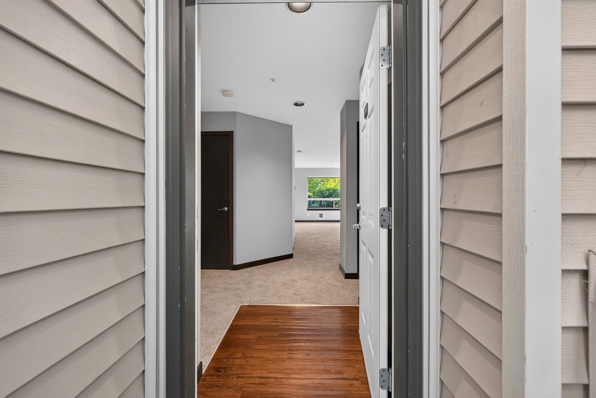 Open doorway to a bright hallway with beige carpet, wood floor, and a window at the far end.