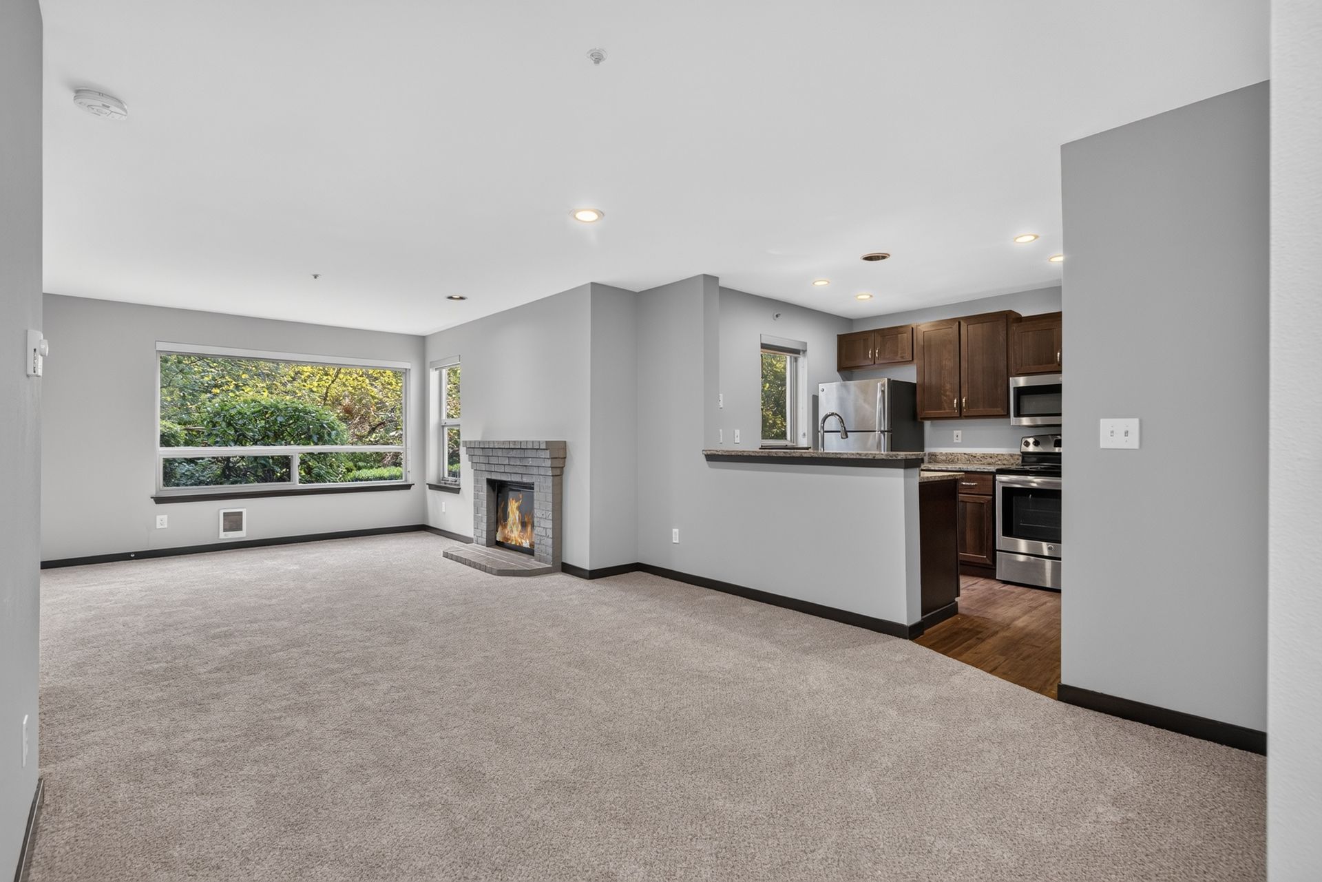 Empty carpeted living room with fireplace and open kitchen, bright window view