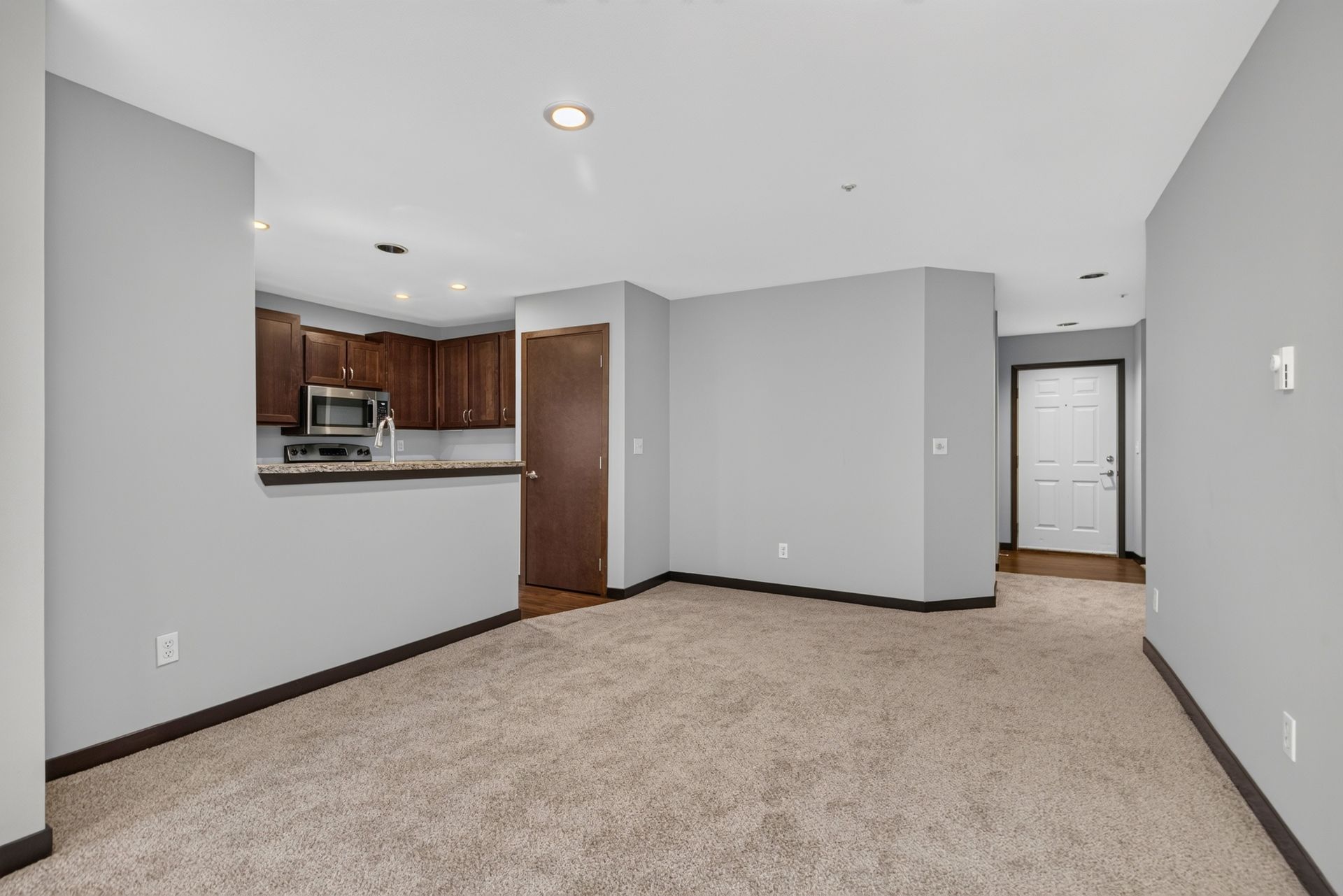 Empty carpeted living room with gray walls, open kitchen, and doorway to hallway