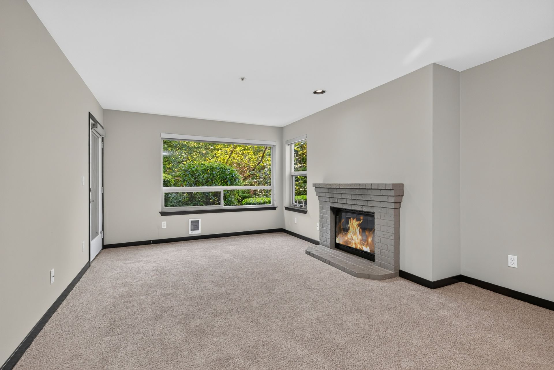 Empty carpeted living room with large window and lit corner fireplace