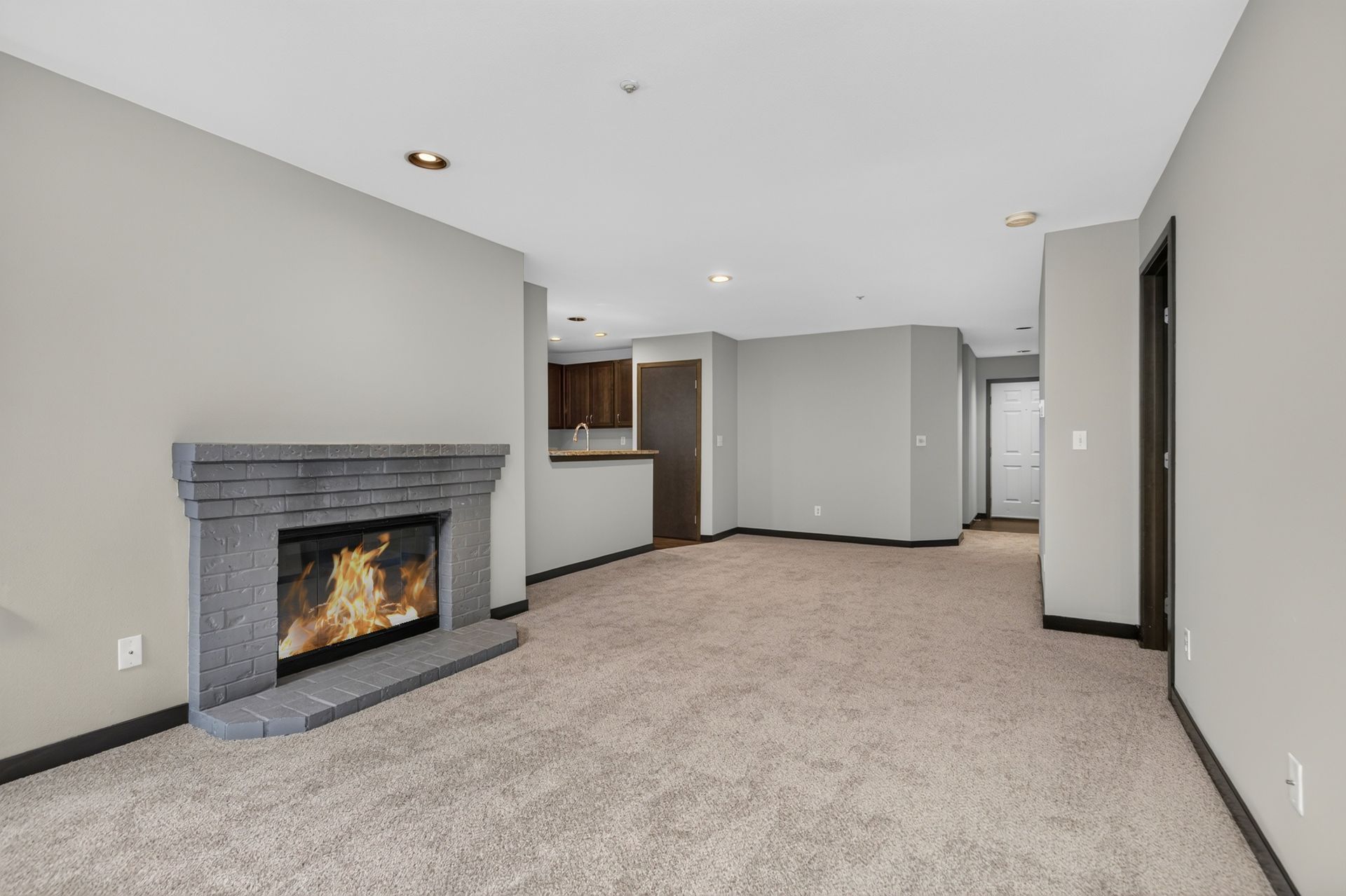 Empty carpeted living room with gray fireplace, beige walls, and open kitchen area.