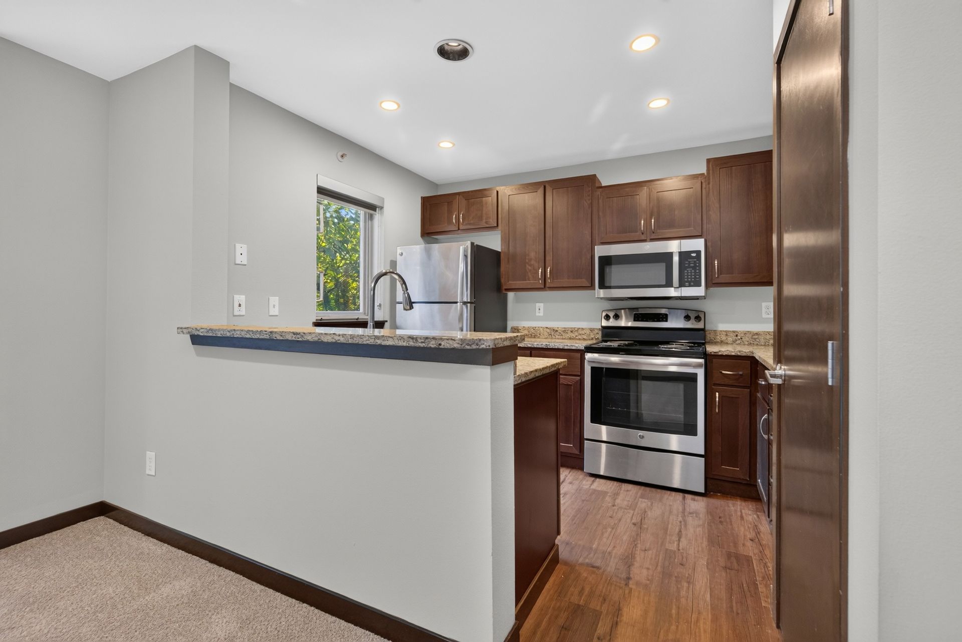 Compact kitchen with wood cabinets, stainless appliances, granite counter, and white walls with wood floor