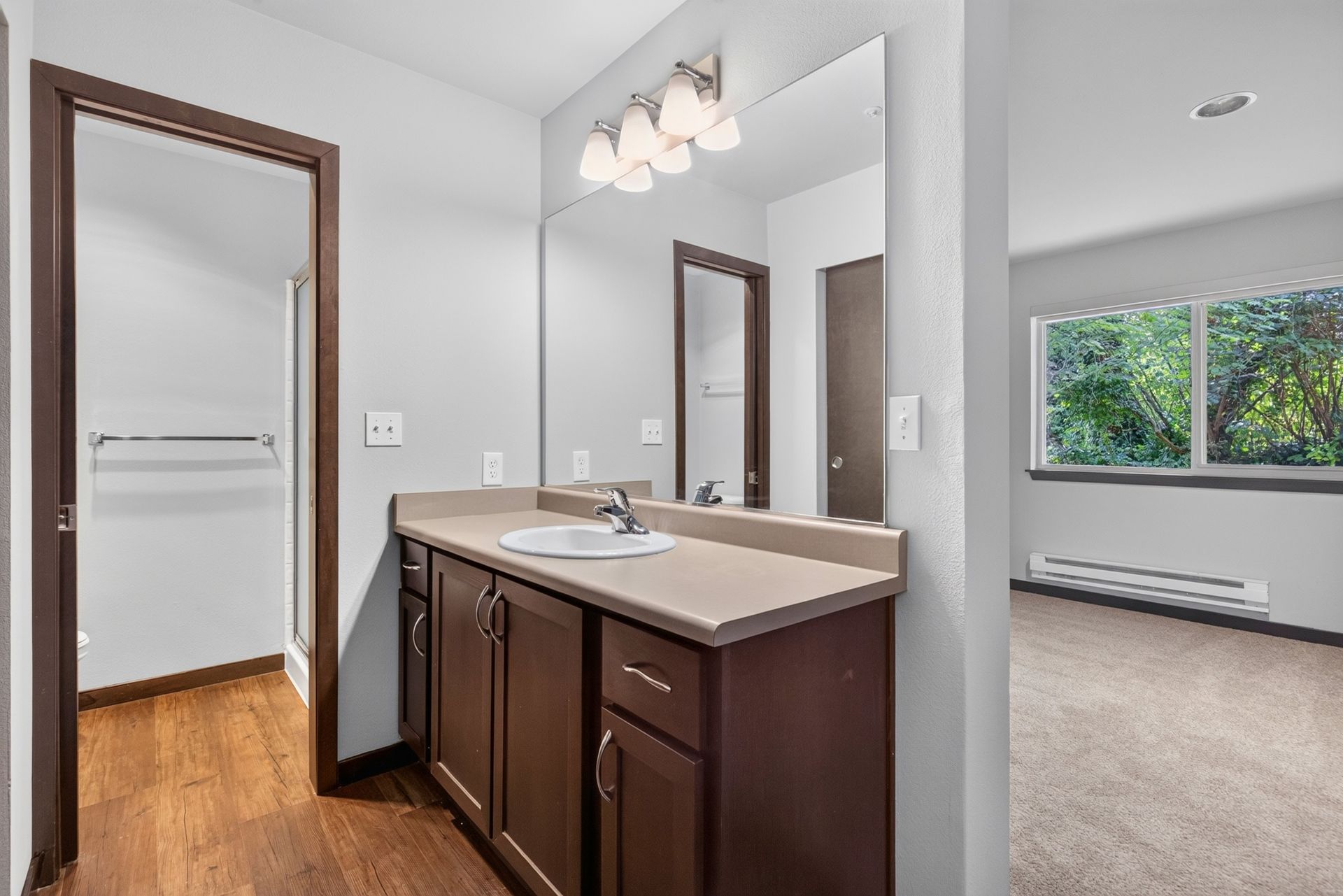 Bathroom vanity with mirror beside a carpeted room and window, showing a doorway to a hall