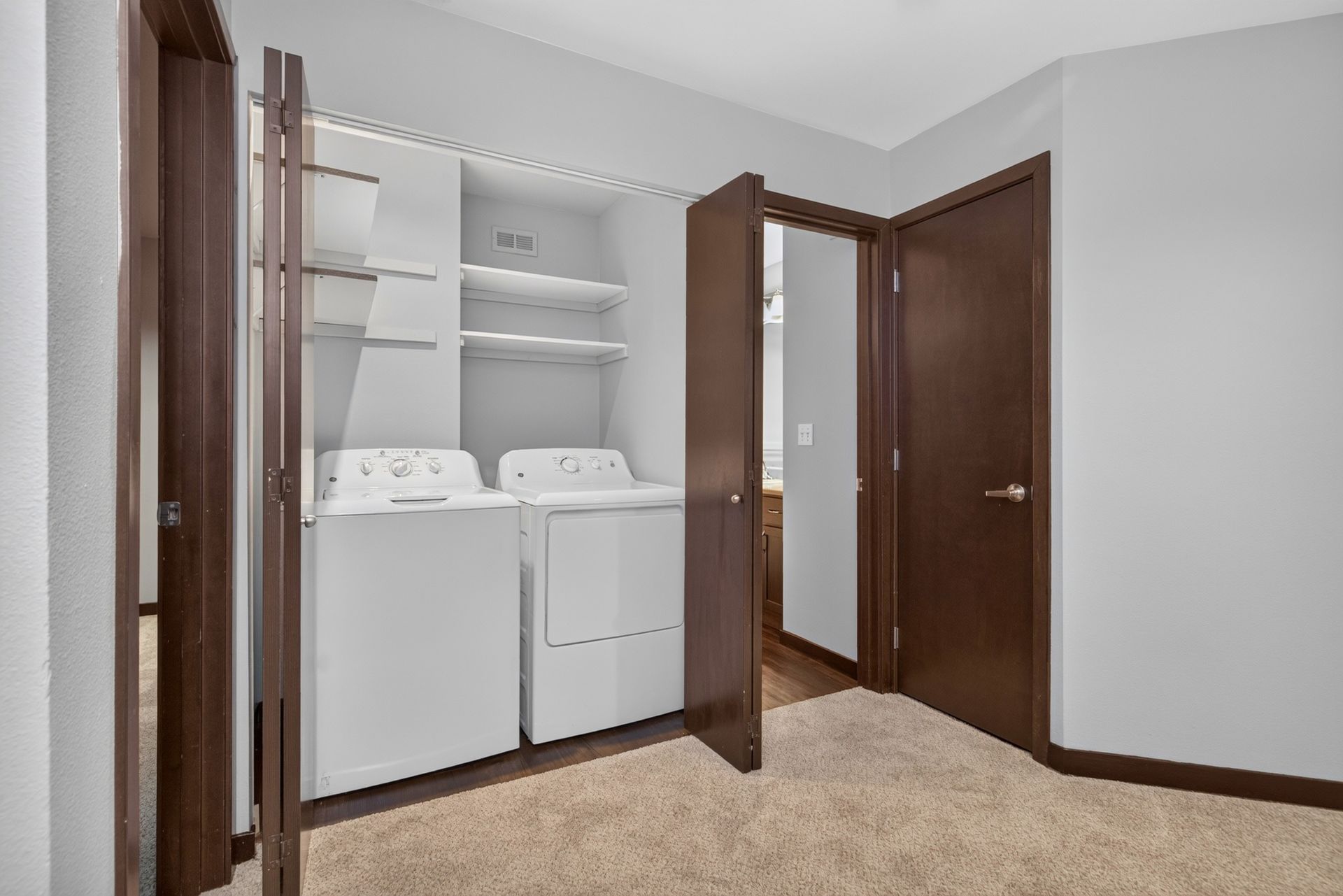 Laundry closet with stacked shelves, white washer and dryer, and brown double doors in a beige room