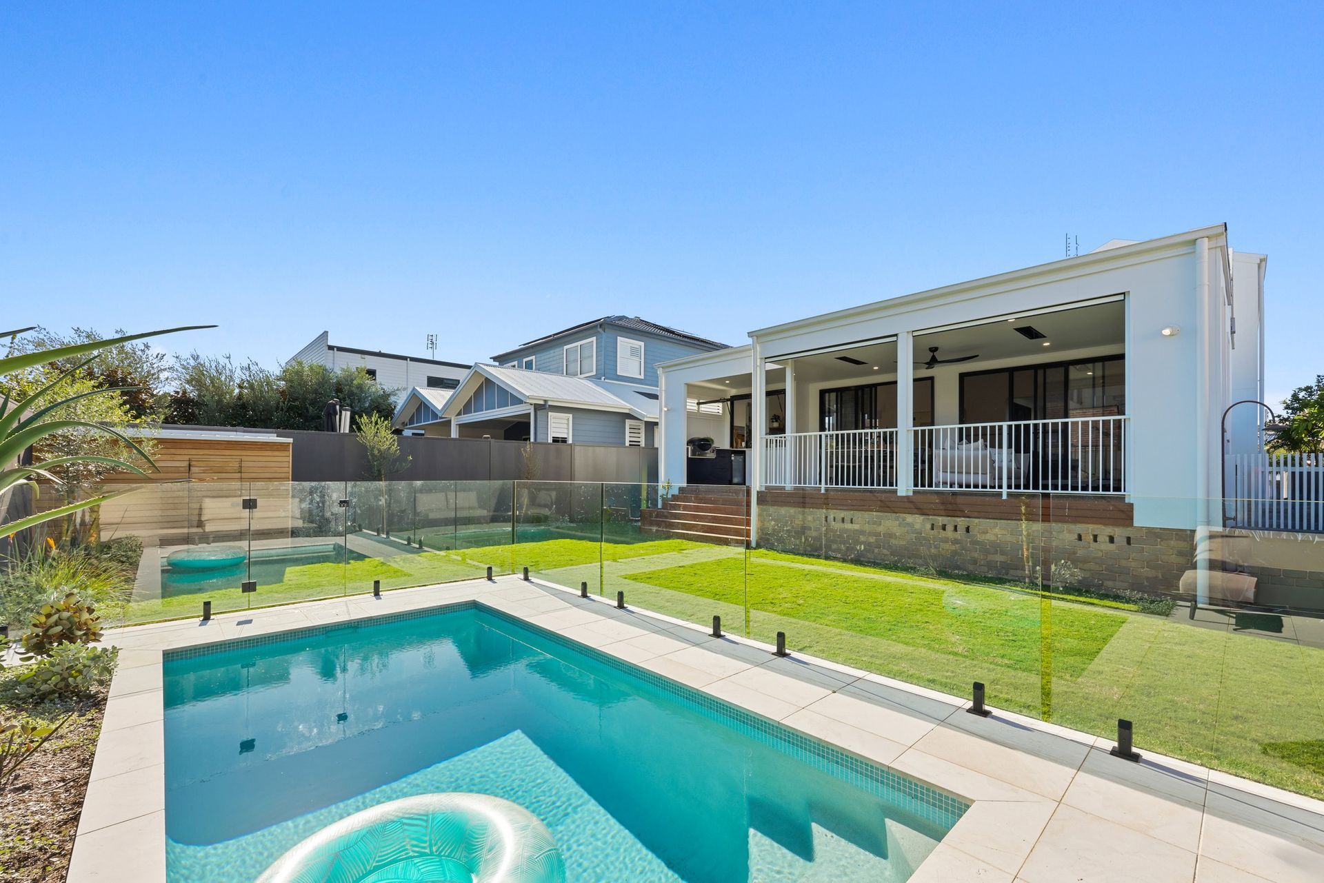 Glass-fenced swimming pool in front of a covered deck, with neighbouring homes behind