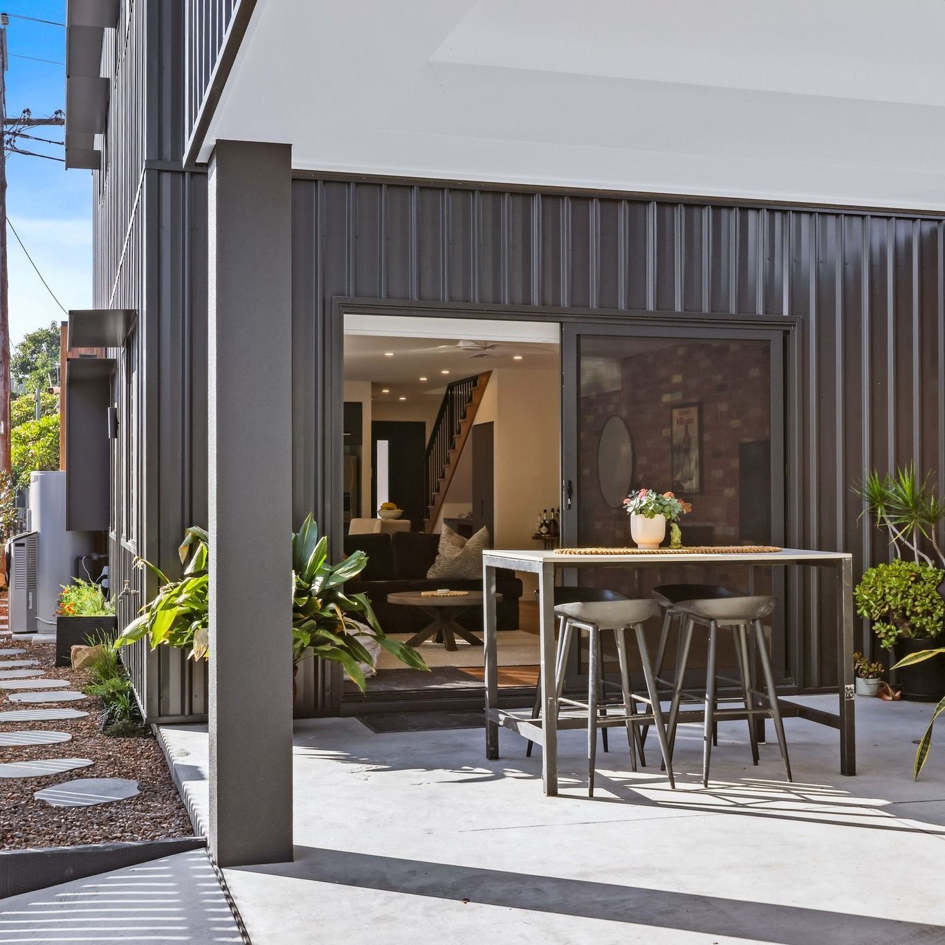 Outdoor dining area with bar stools beside dark metal cladding and a stepping-stone path