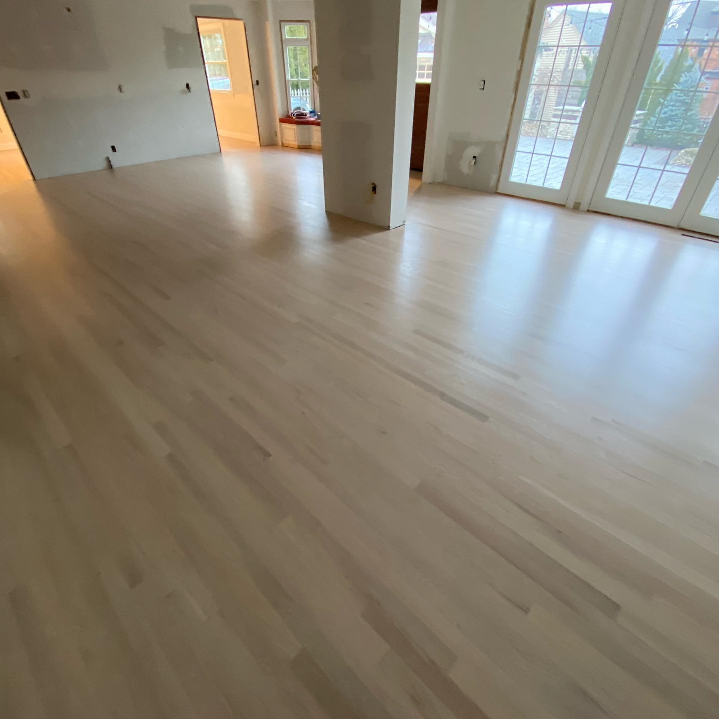 Empty bright living room with light wood floors, large windows, and an open doorway to a kitchen.