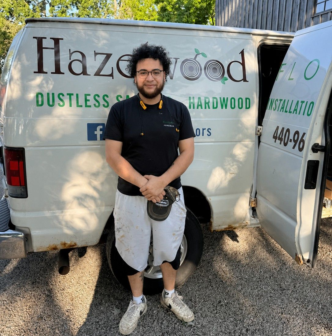 Man standing in front of a Hazeldwood Dustless Hardwood service van, holding a tool, outdoors on gravel.