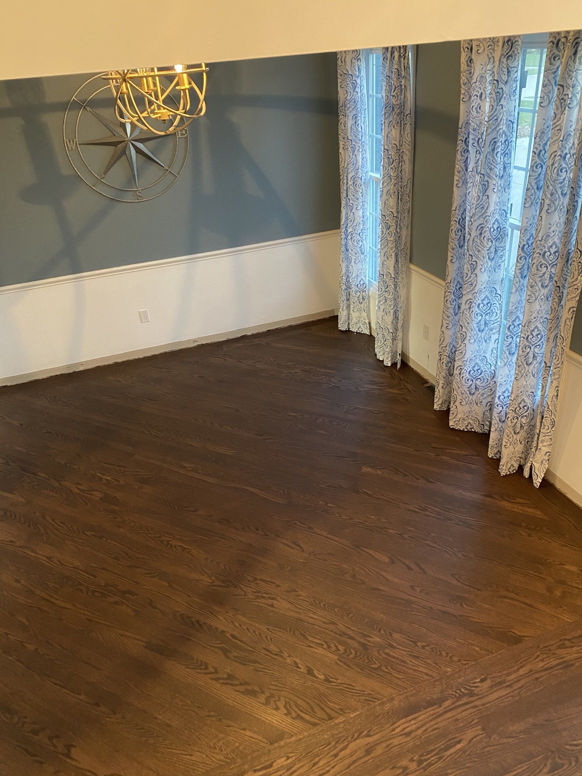Empty room with dark wood floor, blue-gray walls, and sheer curtains by a window.