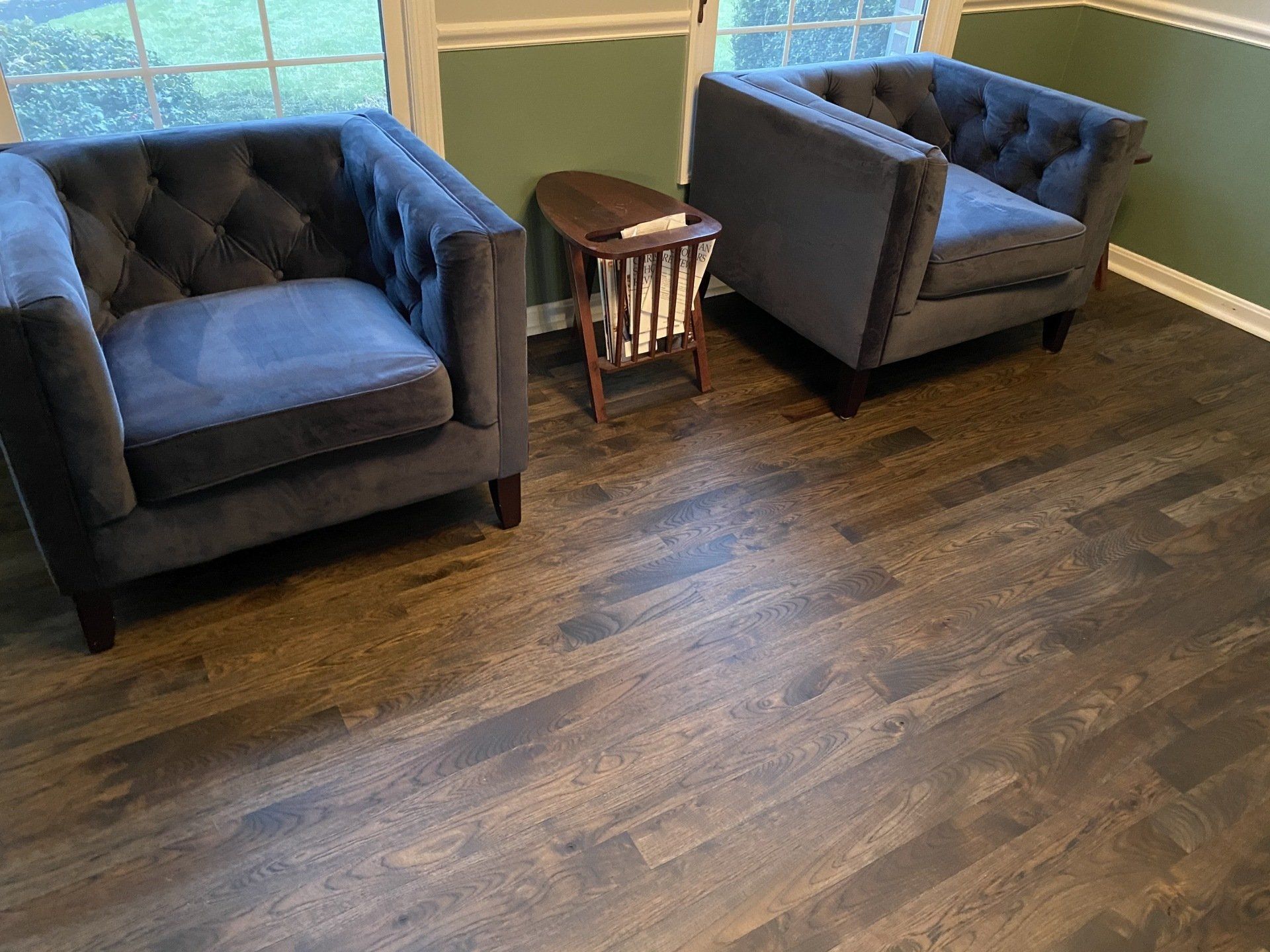 Two blue armchairs with a small wooden side table on a hardwood floor by a window.