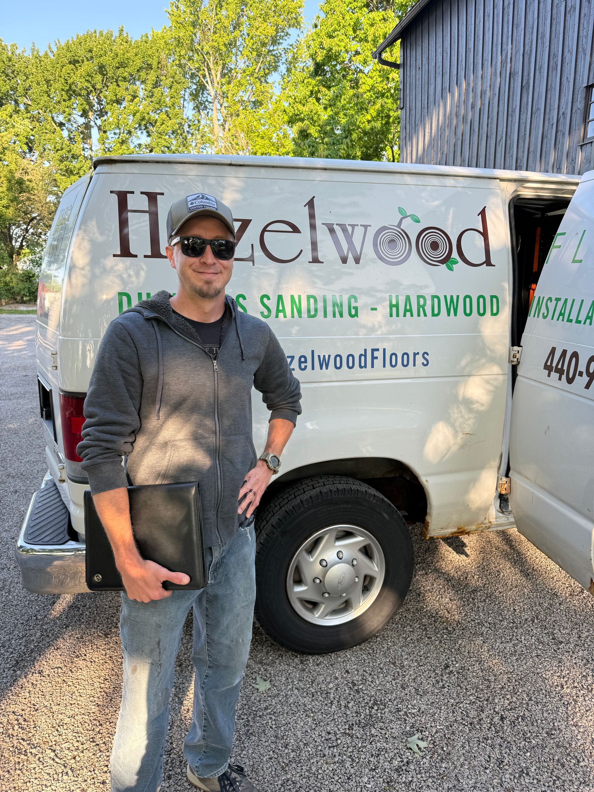 Man standing beside a Hazelwood sanding hardwood work van on a gravel driveway, holding a tool.