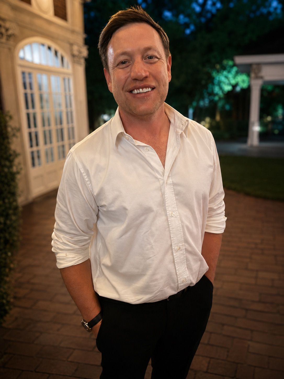 Smiling person in a white shirt and black pants standing outdoors at night near a lit building