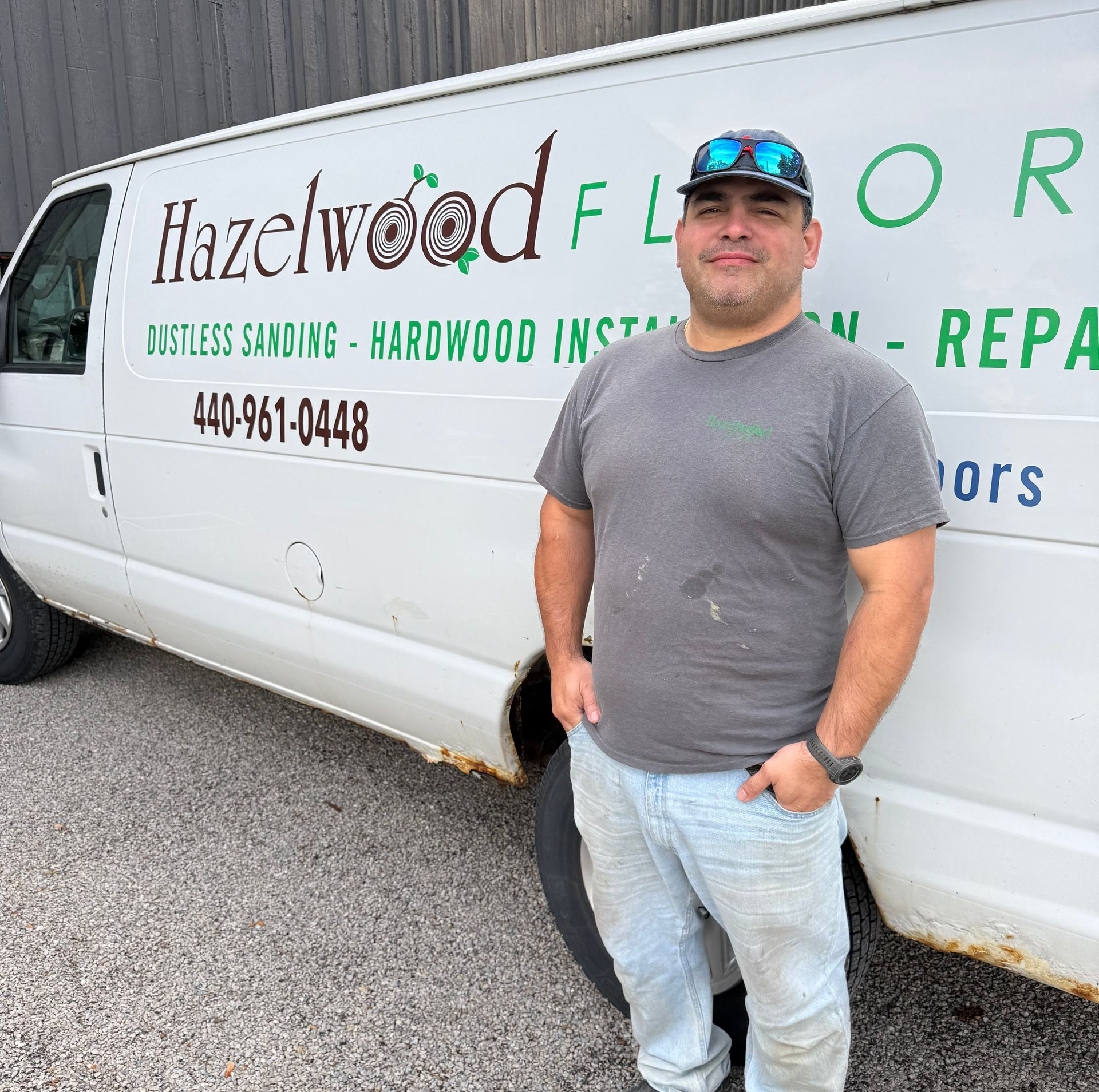 Man standing beside a Hazelwood Floors van in a parking lot, wearing a cap, gray shirt, and jeans.