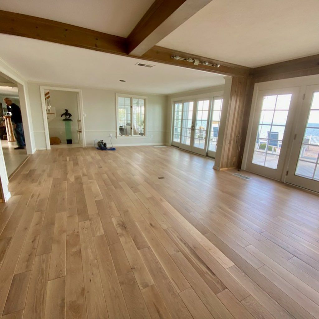Spacious empty living room with light wood floors, exposed beams, and large windows and French doors.