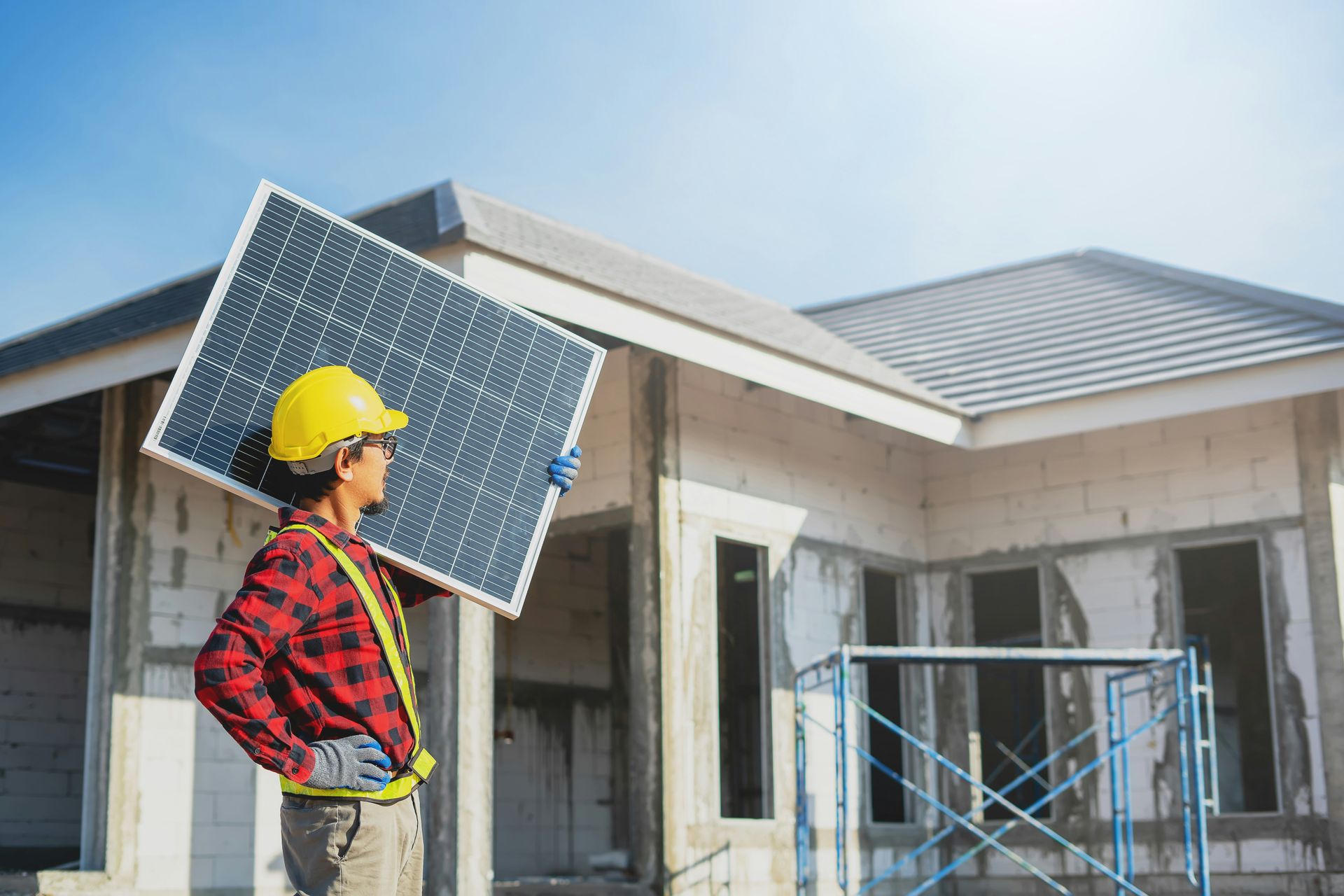 A worker in a yellow hard hat and red plaid shirt carries a solar panel in front of a house under construction.