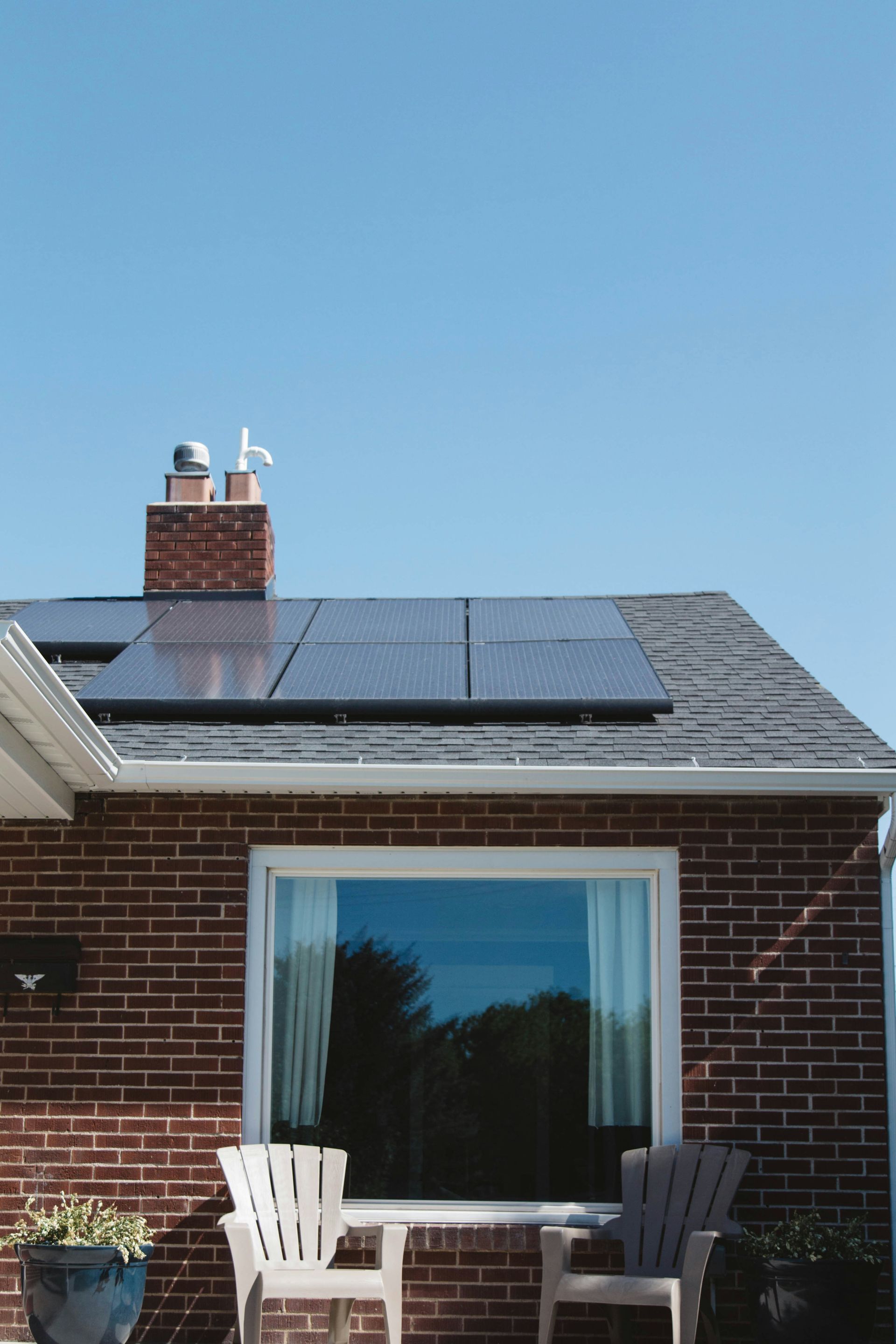A single-story brick house with a solar panel array on the roof, a central window, and two patio chairs in front.
