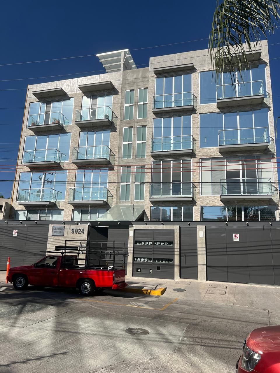 Edificio moderno de hormigón con grandes ventanales y balcones, camioneta roja estacionada al frente.