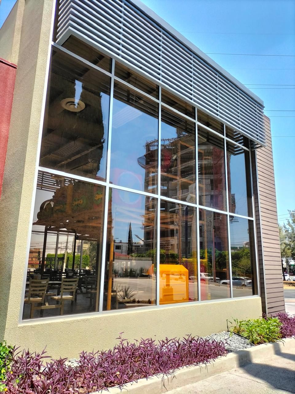 Grandes ventanales reflejan un edificio y un interior con plantas exteriores debajo y una sombrilla de metal arriba.