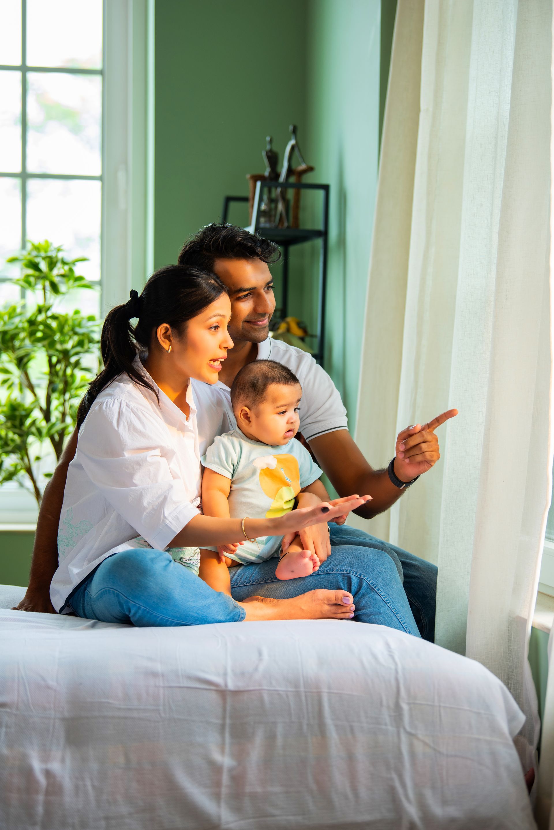 Family with baby looking out a window, pointing, sitting on a bed. Bright sunlight.