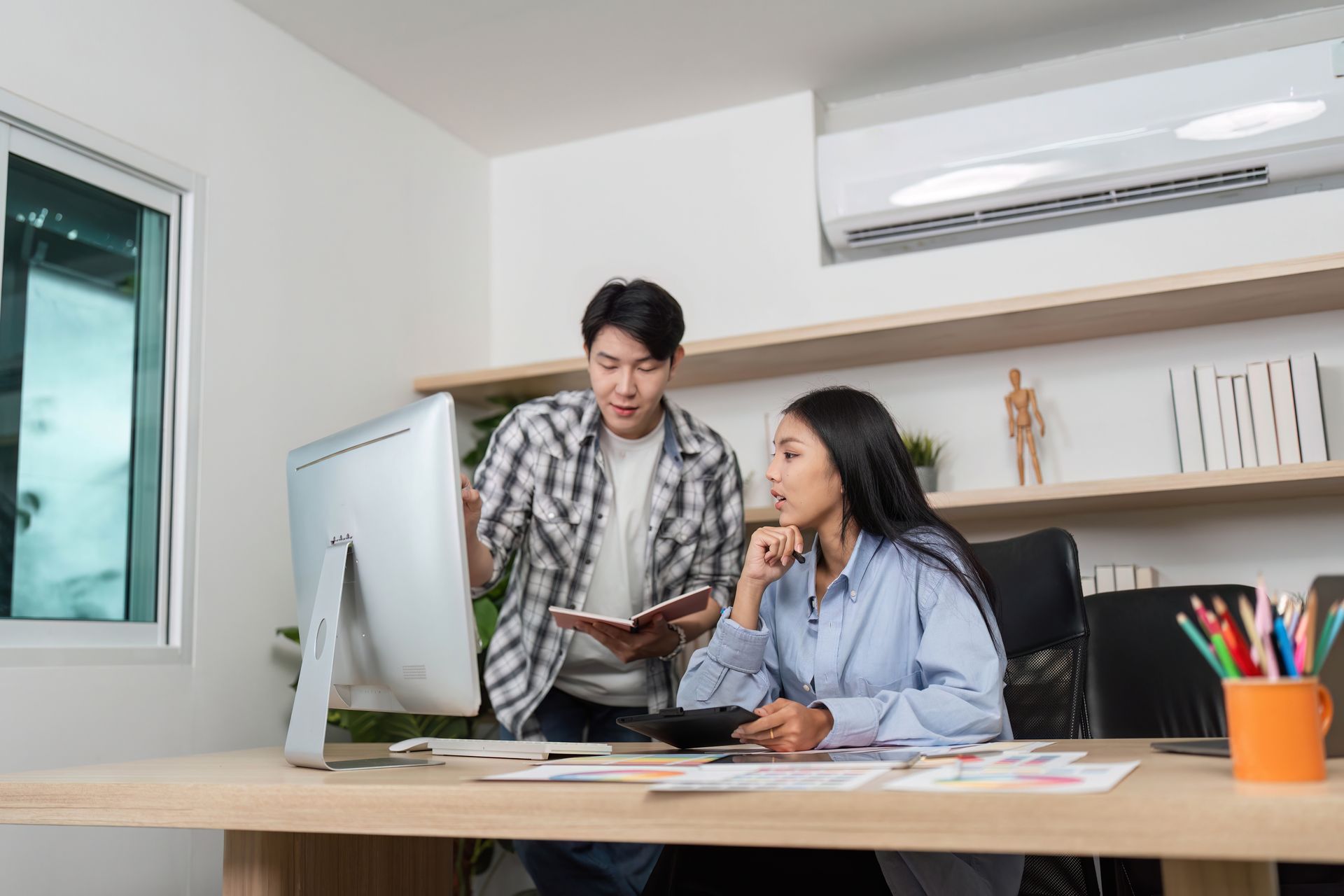 Two people, one standing, the other seated, looking at a computer monitor in an office.