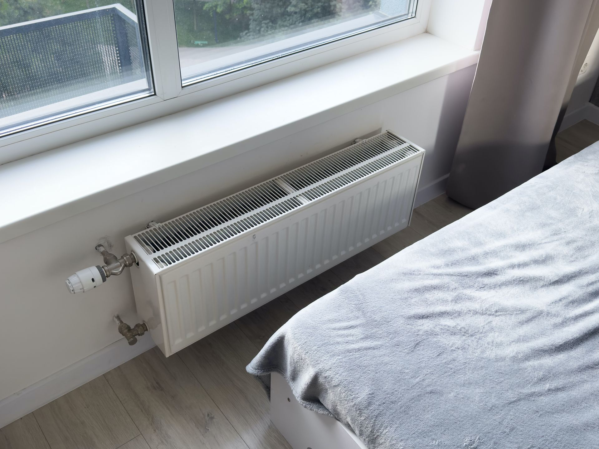 White radiator below a window next to a bed in a room.