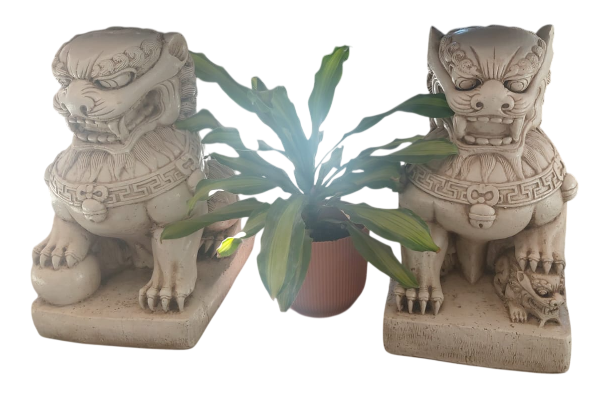 Dos estatuas de leones guardianes de piedra flanquean una planta verde en maceta sobre una superficie blanca.