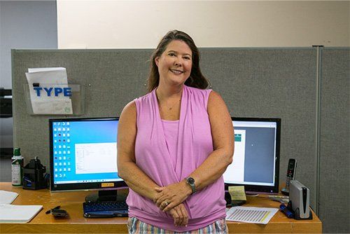 Woman in pink top smiles in front of a desk with two computer monitors in an office.