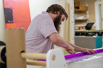 Man with beard cutting paper with a purple ruler in a workshop setting.