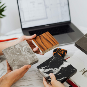 Hands holding three patterned tile samples over a laptop on a desk