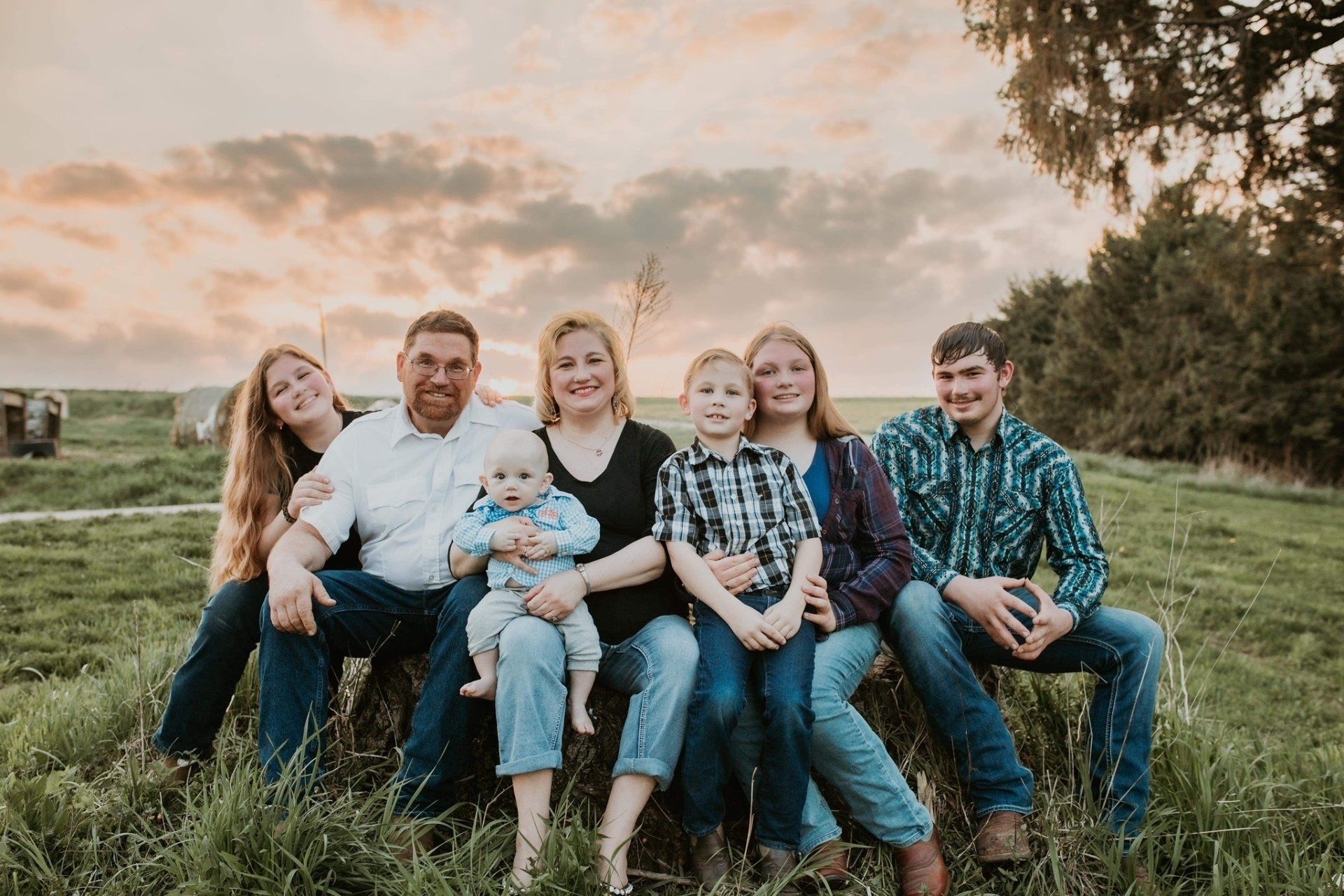Family of seven sitting in a grassy field; cloudy sunset background.