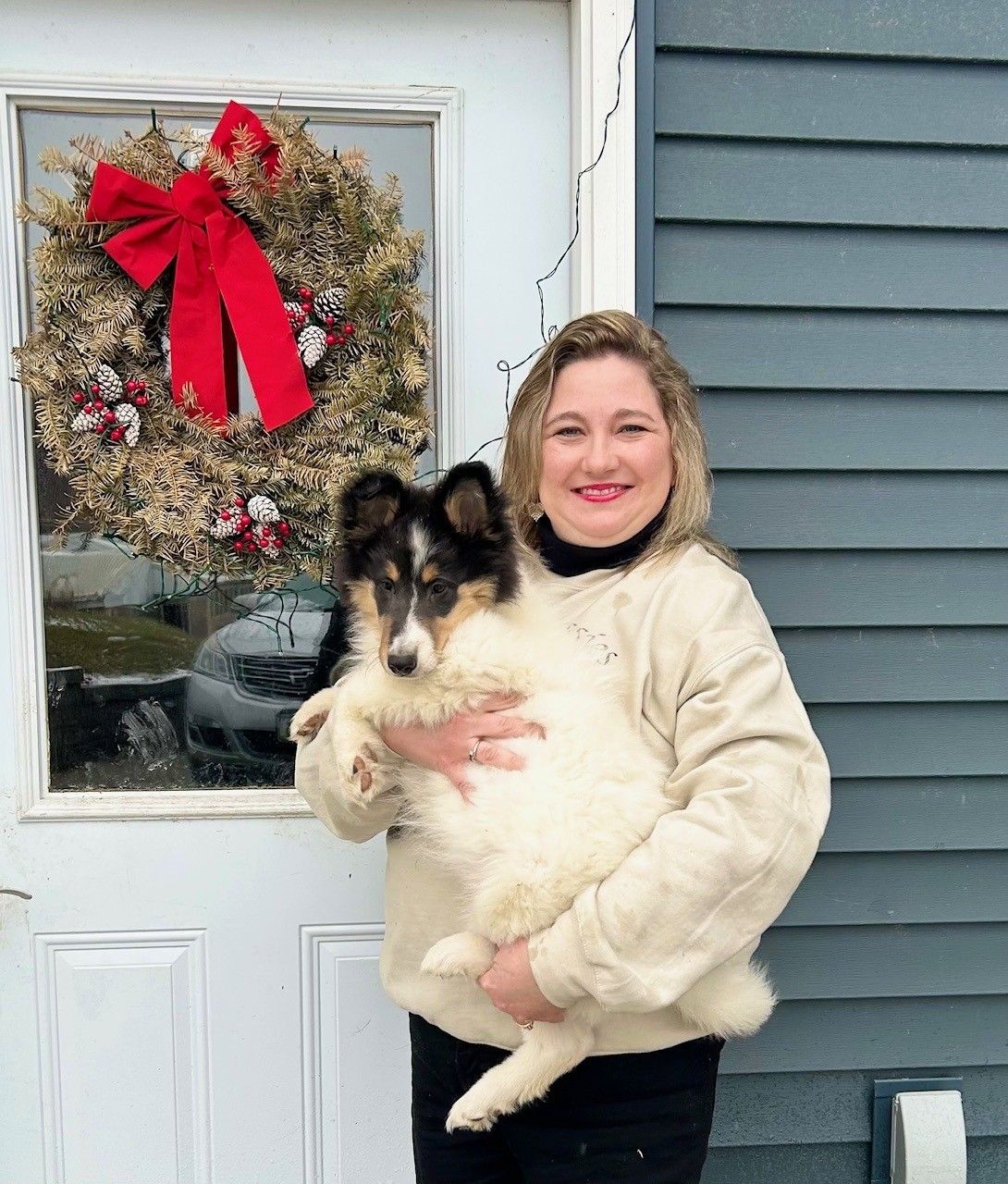 Woman holding a fluffy dog in front of a door with a holiday wreath.