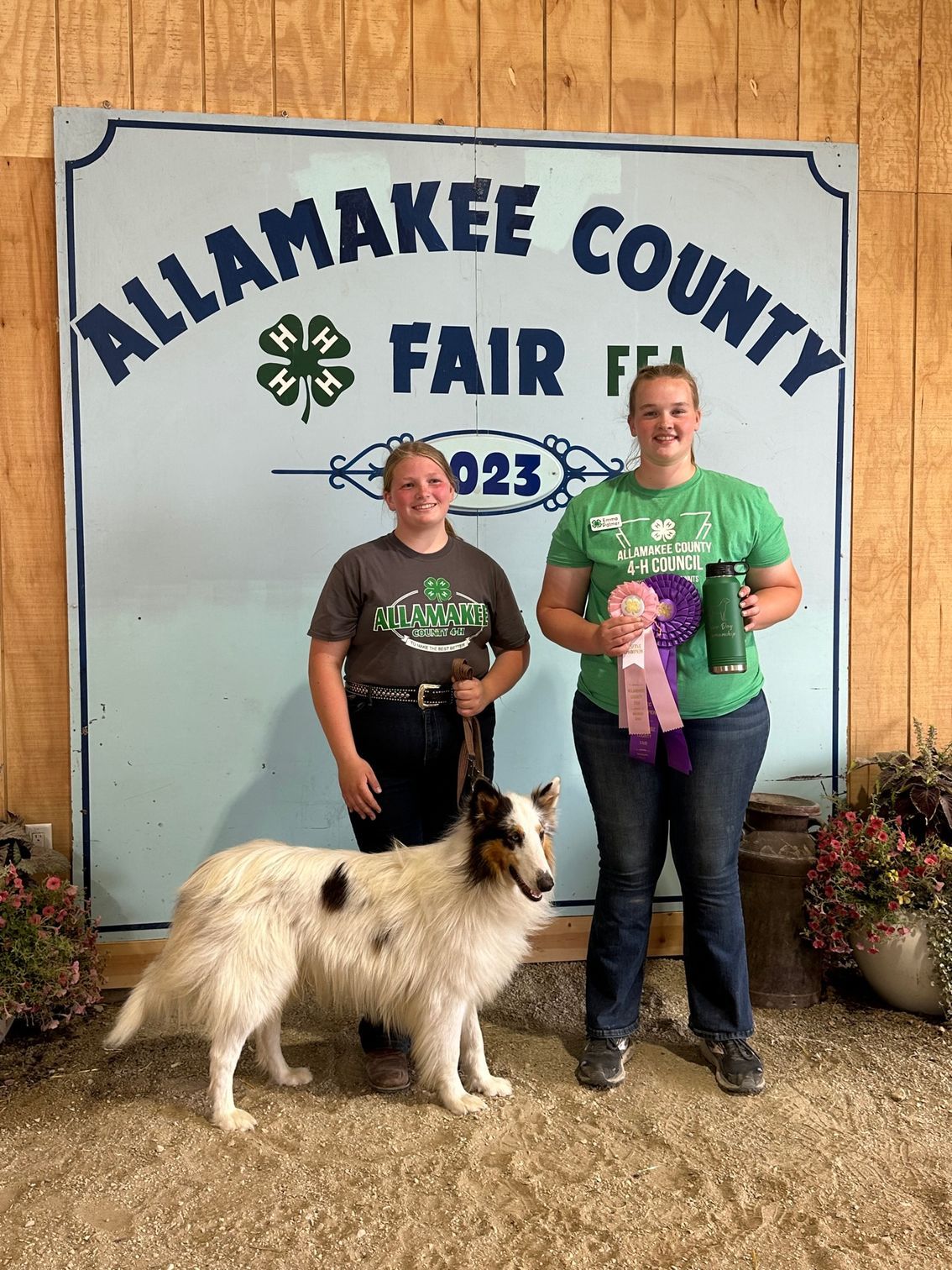 Two people with a dog at Allamakee County Fair. One holds ribbons, the other a leash. A fair sign is in the background.