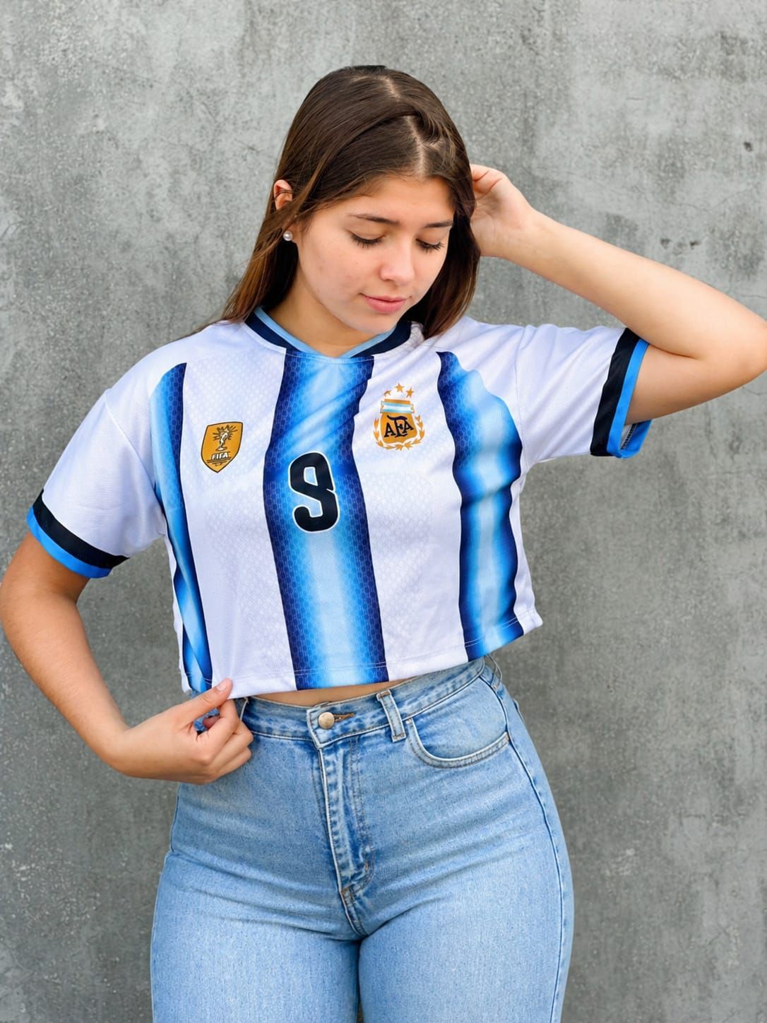 Mujer con camiseta de fútbol de Argentina y vaqueros, posando contra una pared gris.