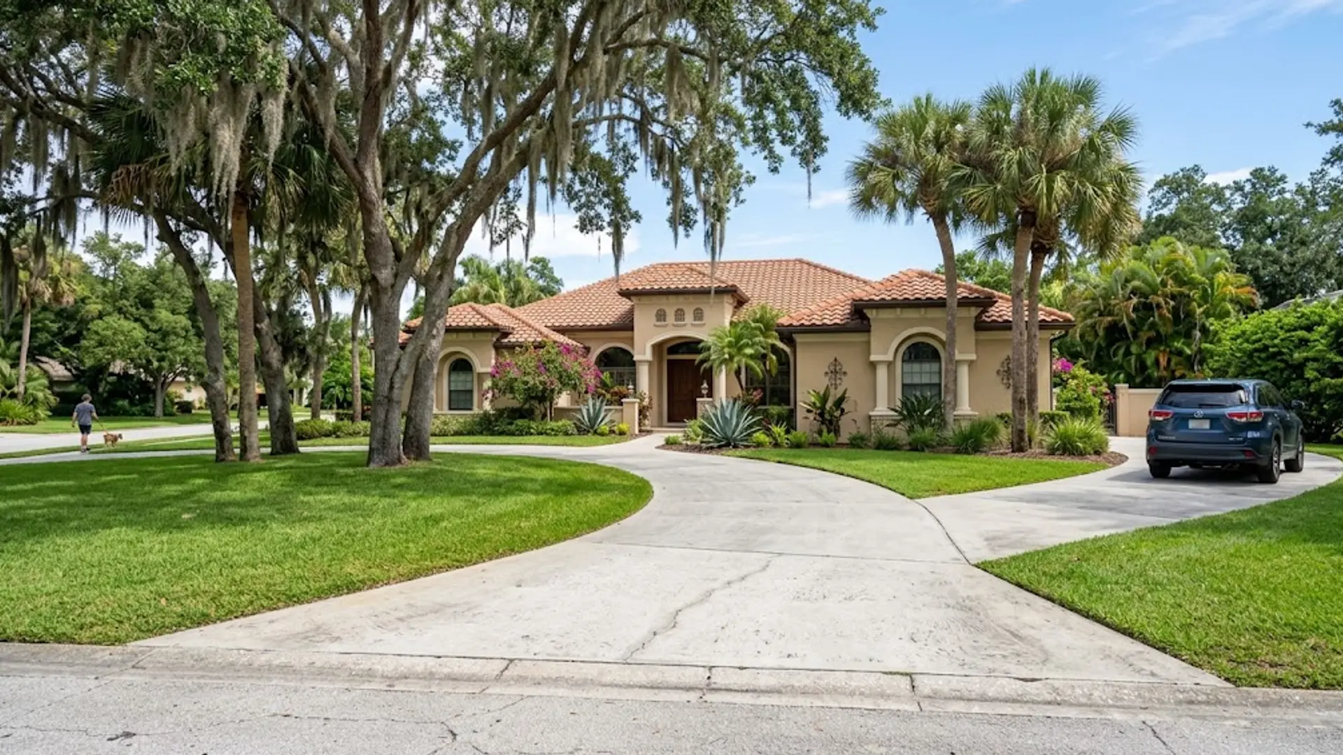 Mediterranean-style house with red tile roof, curved driveway, lawn, and parked SUV under palm trees