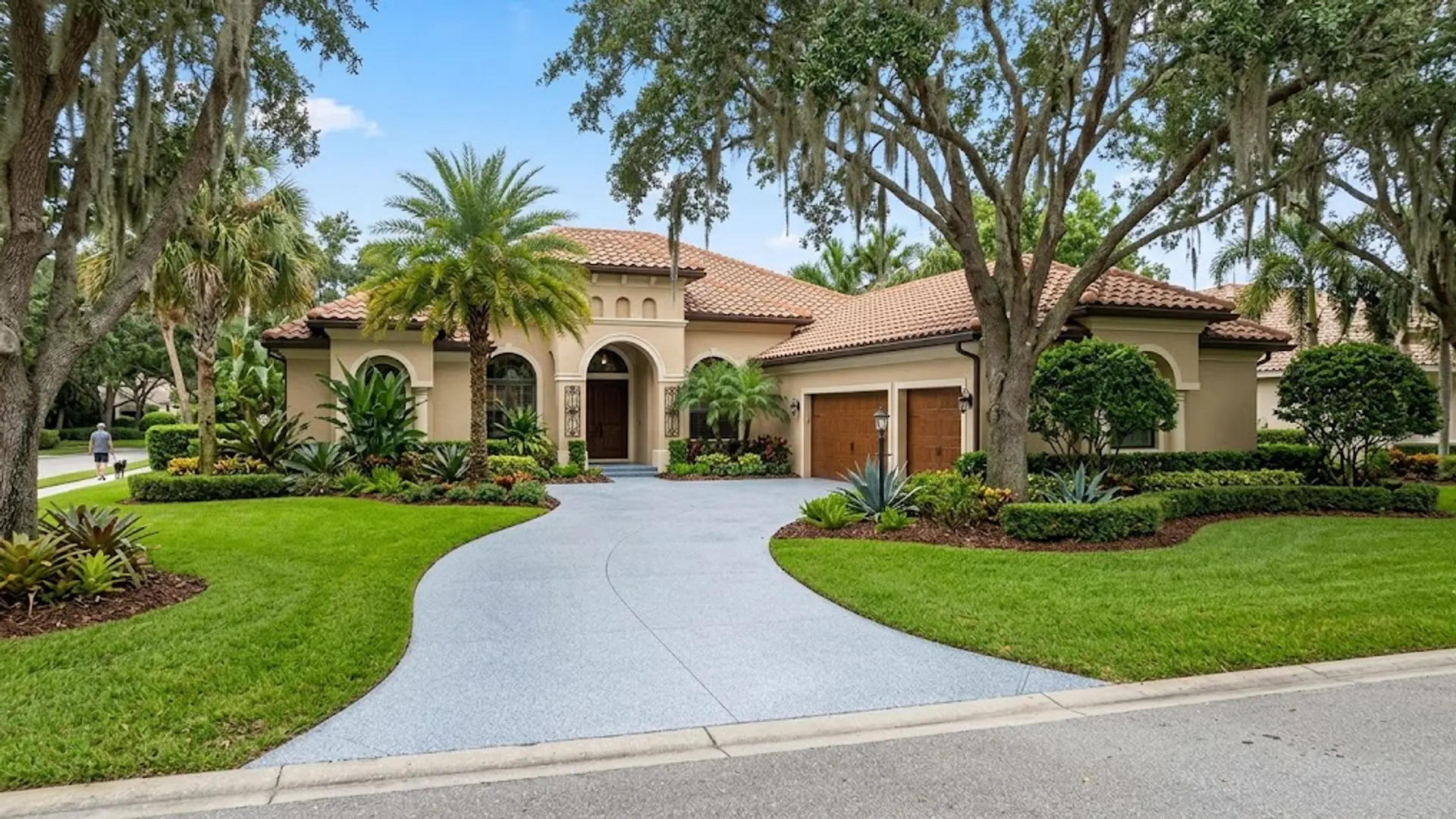 Luxury stucco house with a curved driveway, manicured lawn, and tall trees under a clear sky