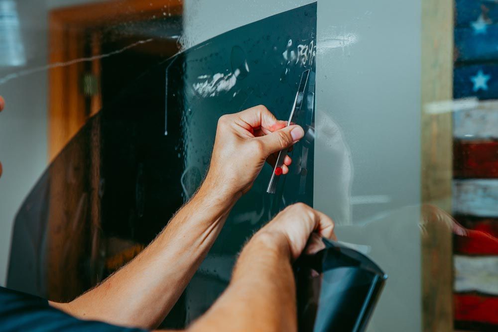A technician using a pen knife to trim window tint film along the edges, preparing it for final installation on a vehicle window.