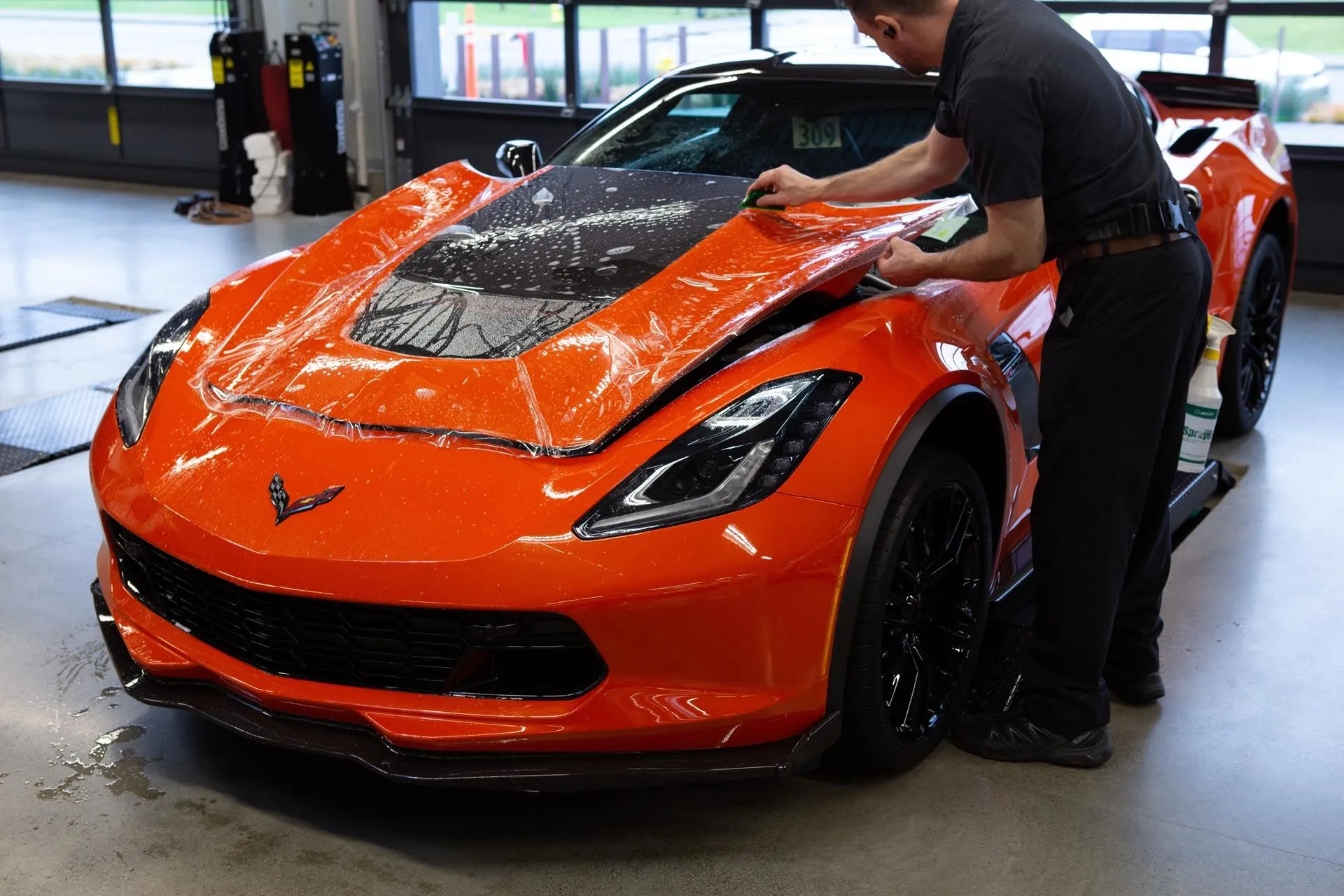A technician is applying paint protection film to the hood of an orange sports car, using a squeegee to smooth the clear film during the installation process.