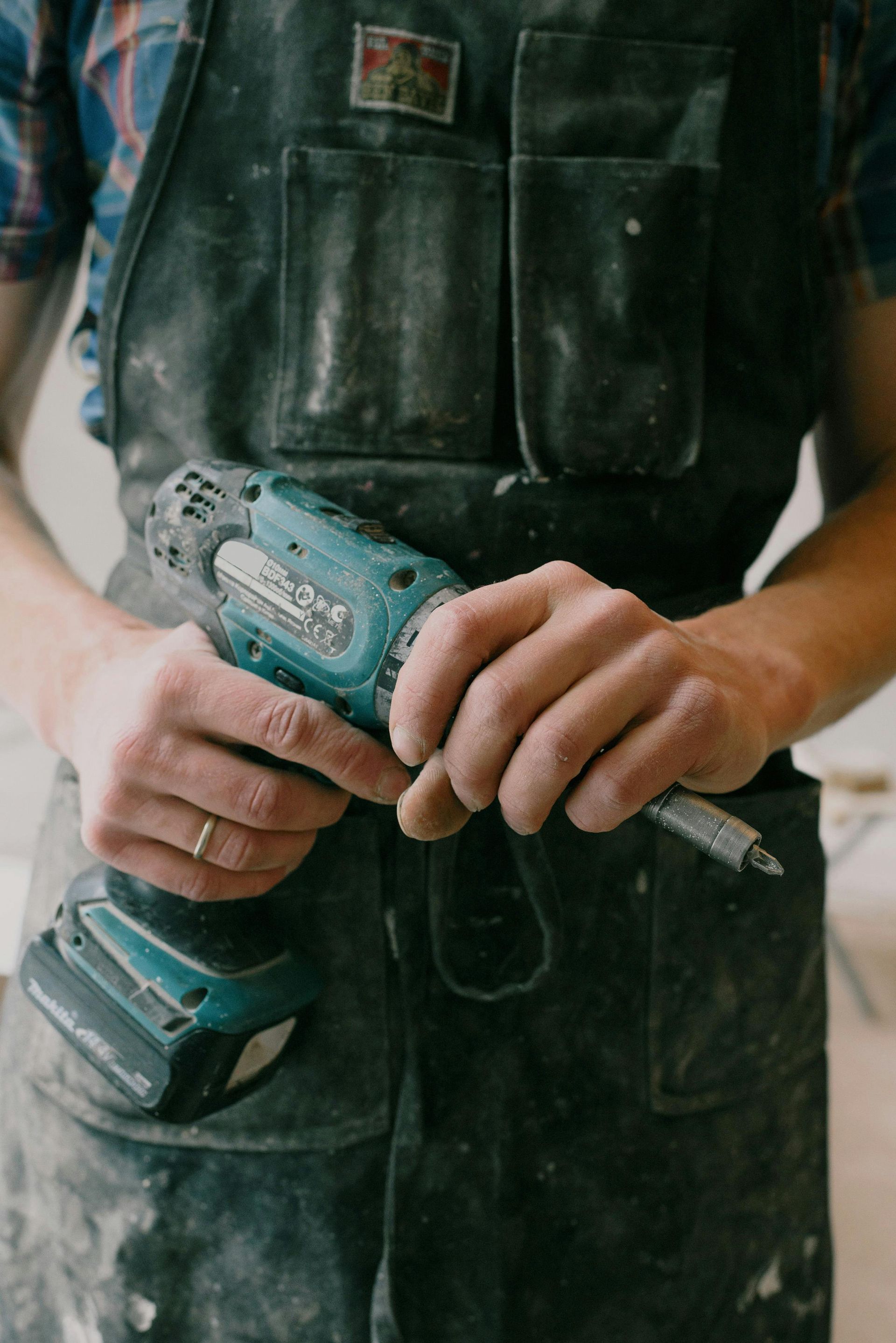 Person in work apron holding a teal and black drill. Indoors.