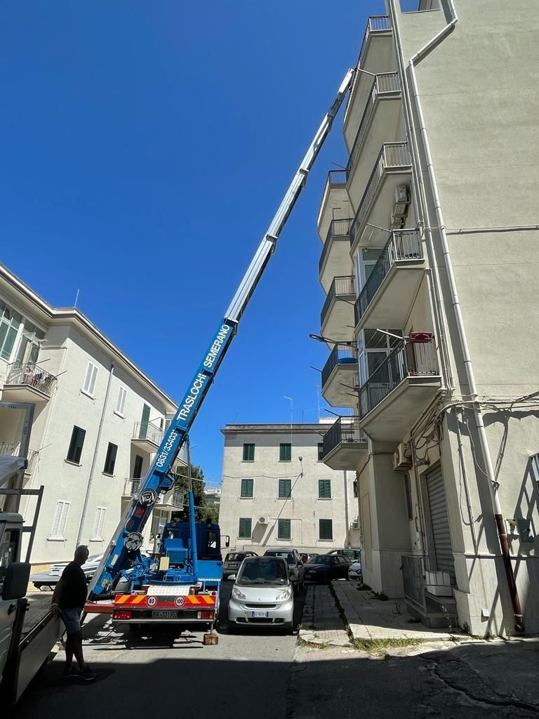 Una lunga gru blu si solleva verso un edificio; una persona è in piedi accanto a un camion; cielo azzurro.