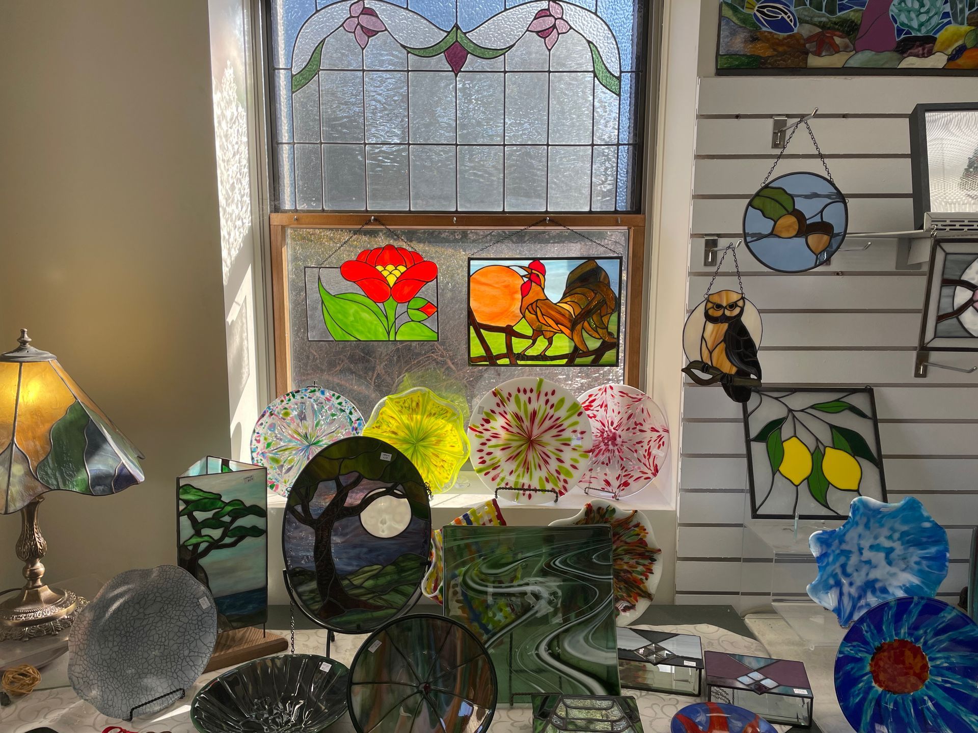 Stained glass art display in front of a window, including floral, animal, and abstract designs.
