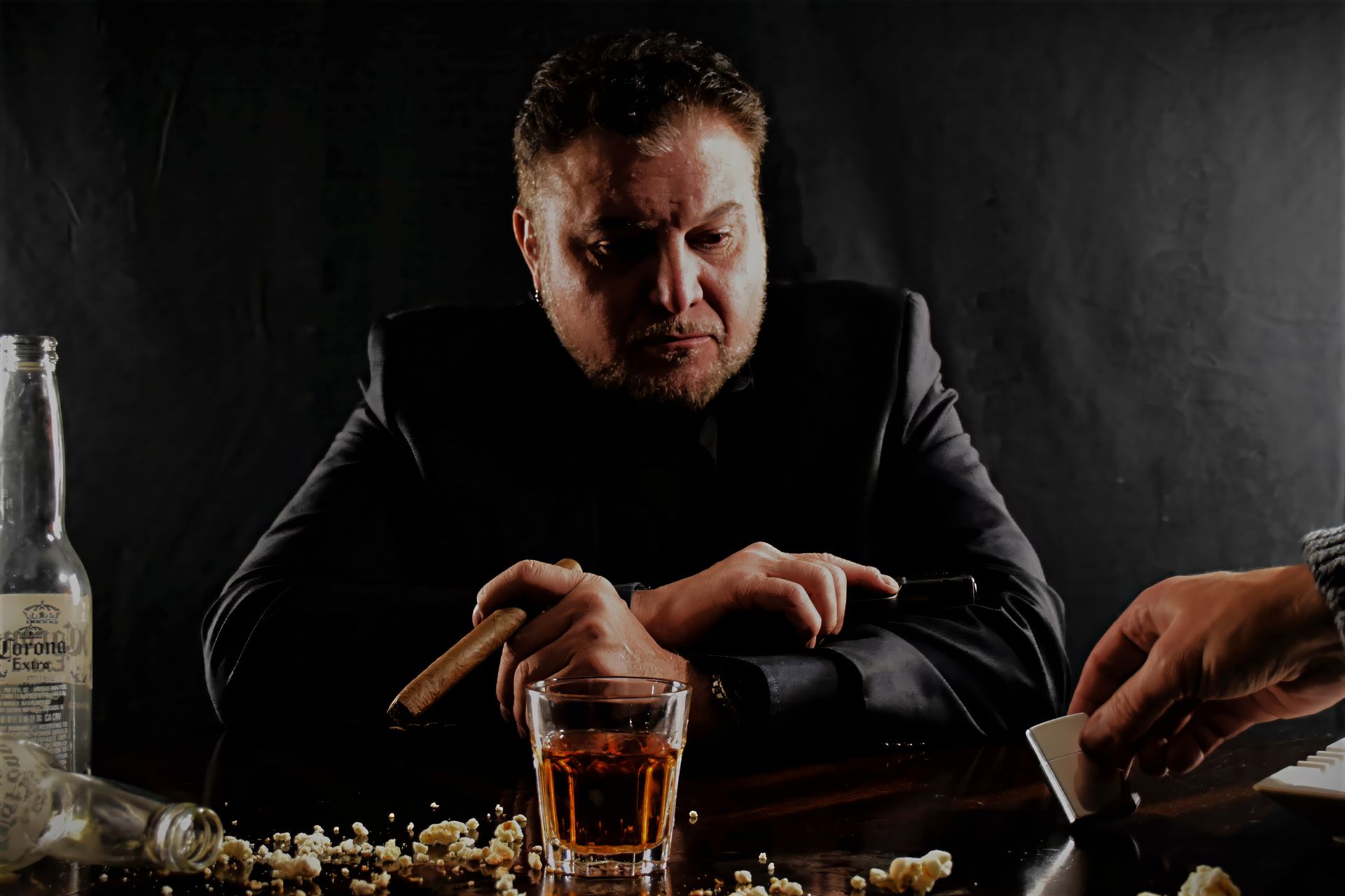 Man in suit smoking cigar, looking at playing cards in dim setting with alcohol.
