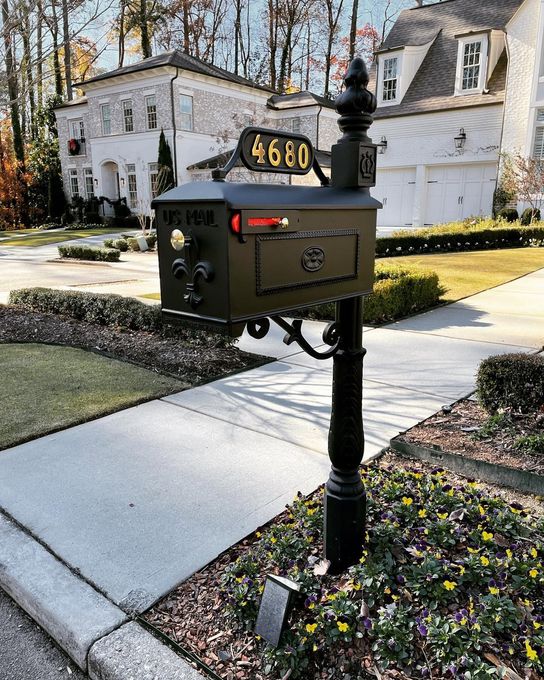 Black mailbox on a post by a sidewalk in a suburban neighborhood, with white houses and autumn trees behind.
