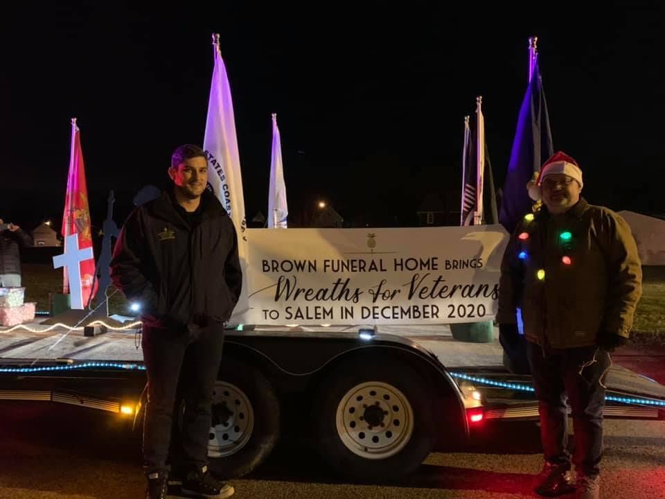 Two men are standing in a cemetery holding a wreath.