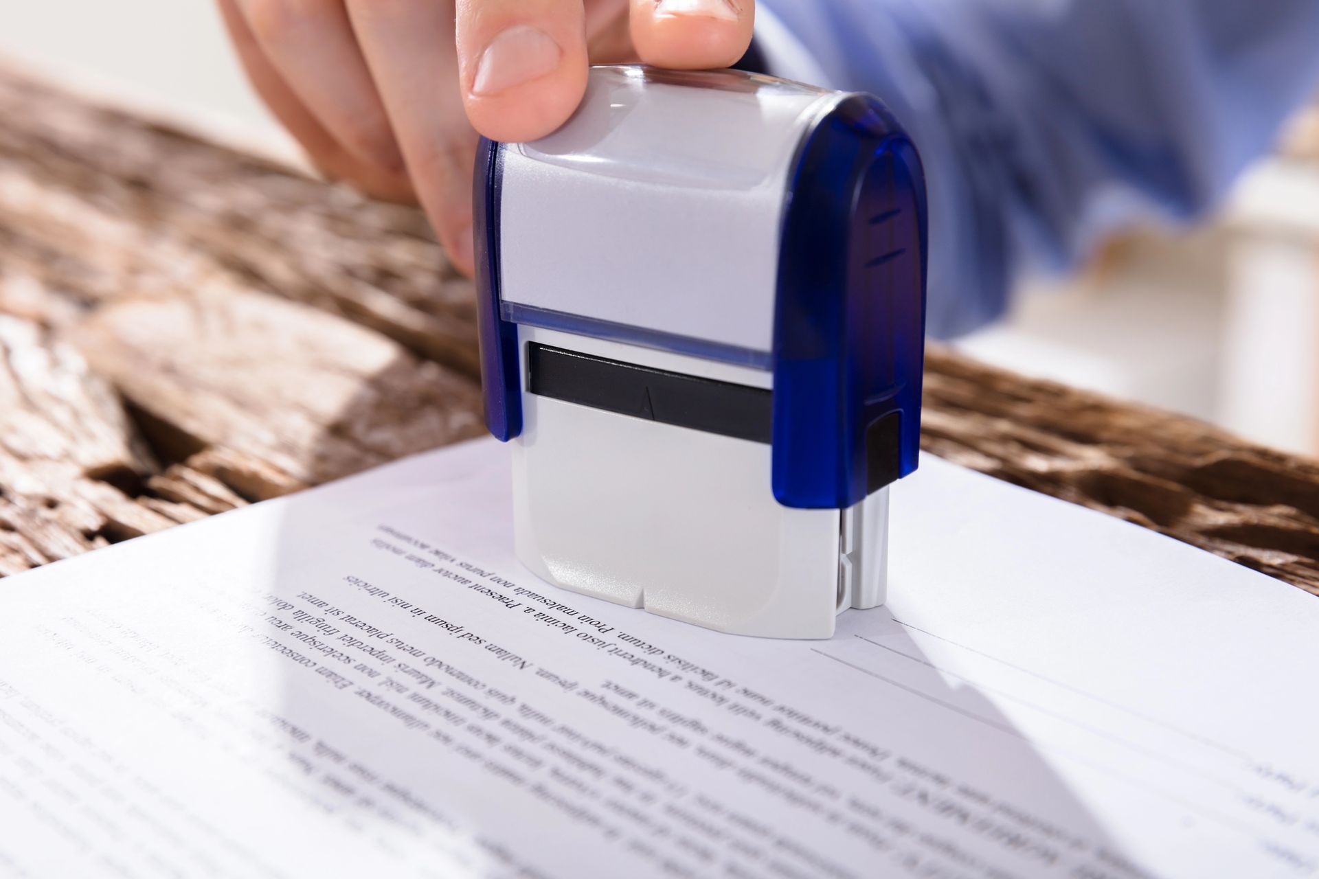 Persona estampando un documento con un sello automático azul y blanco sobre una superficie de madera.
